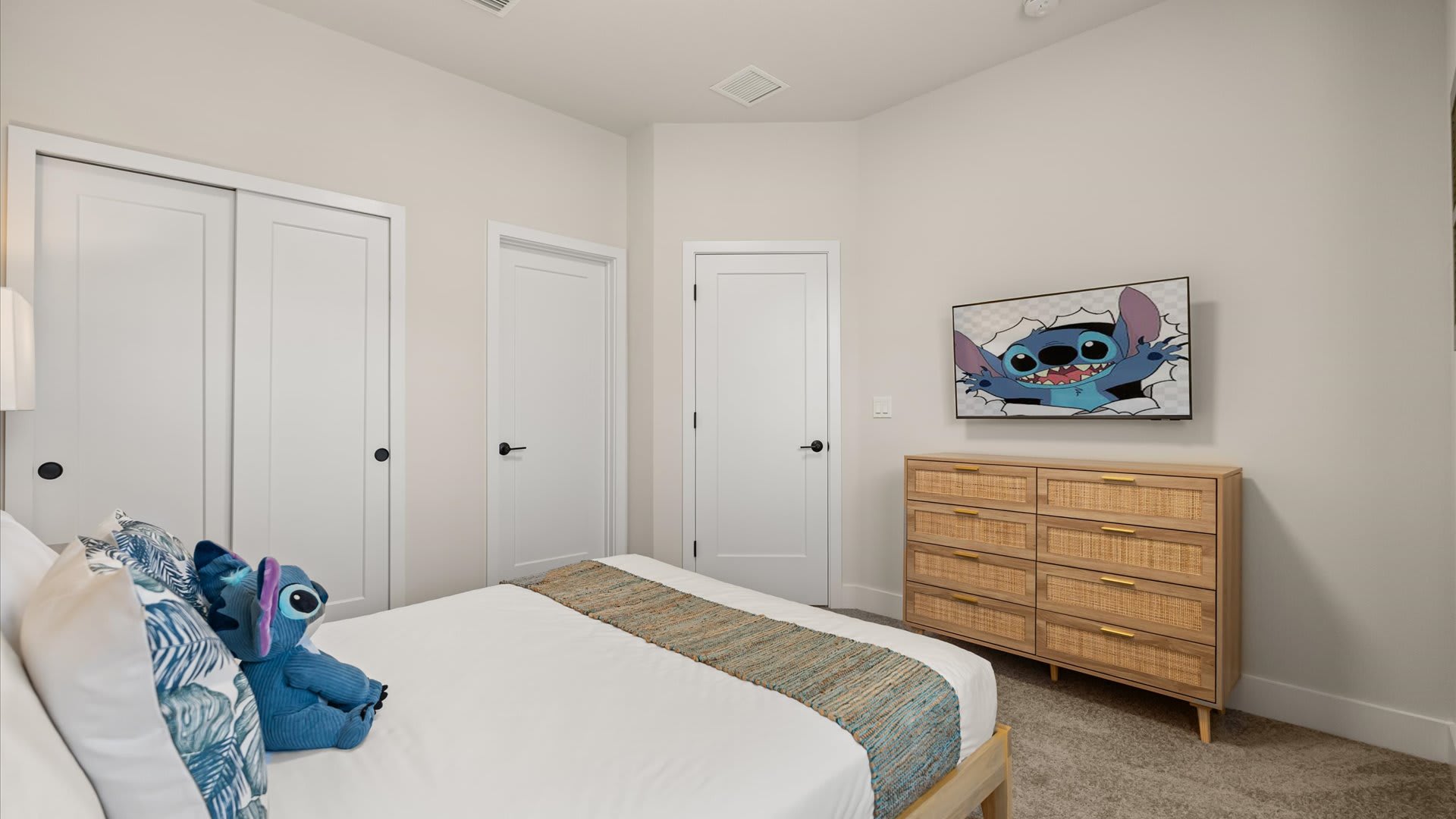 Bright bedroom with light walls, a white bed with blue accents, and a Stitch plush toy. A wooden dresser with rattan drawers and a TV displaying Stitch are prominent.