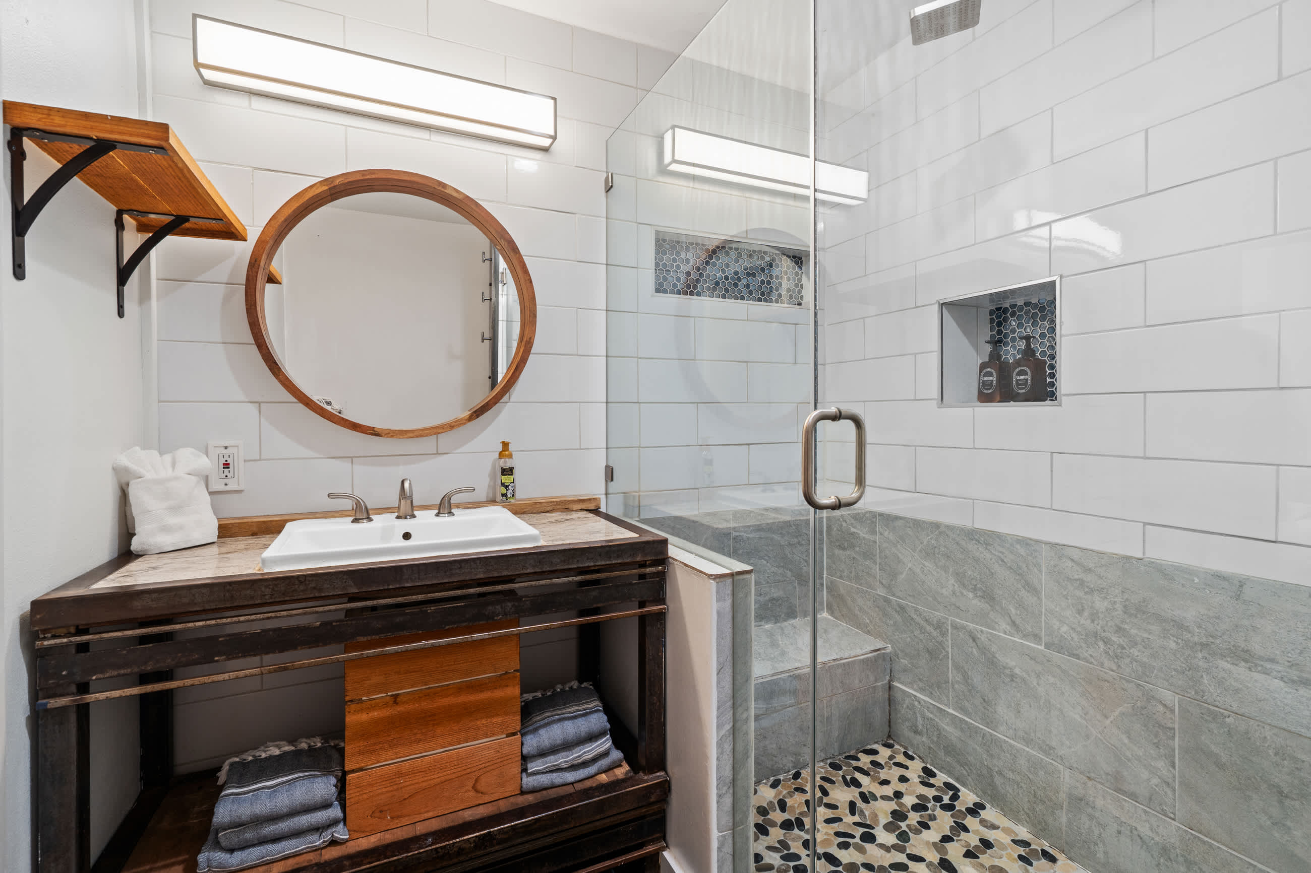 Modern bathroom featuring a dark wood vanity with a white sink, a round wooden mirror, and white subway tile walls. The walk-in shower has a glass enclosure, pebble floor, and a built-in niche with decorative tiles.