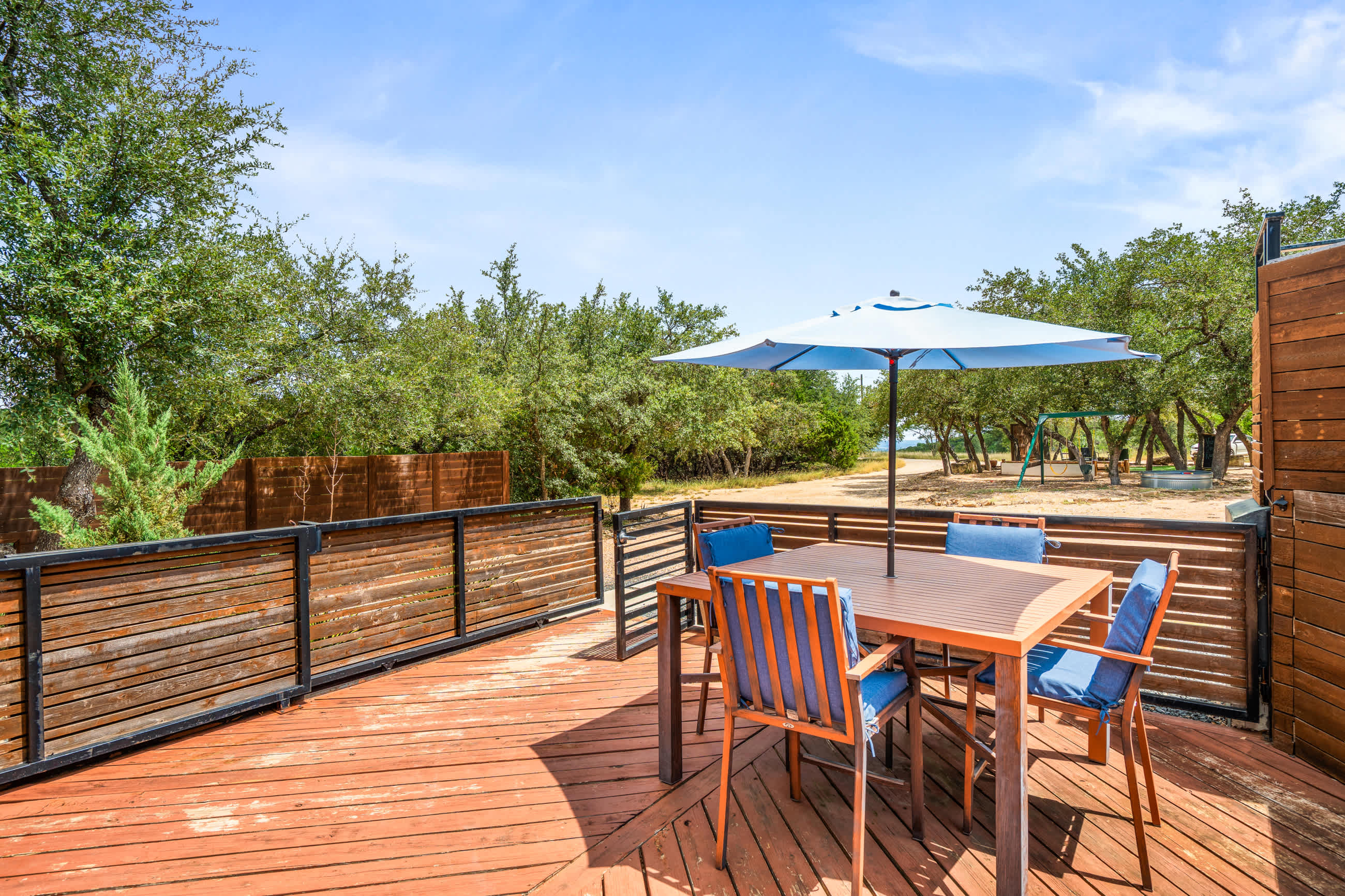 Spacious wooden deck featuring an outdoor dining table with blue chairs and a matching umbrella, enclosed by a slatted wooden fence. The background reveals a natural landscape with trees and a distant play area.
