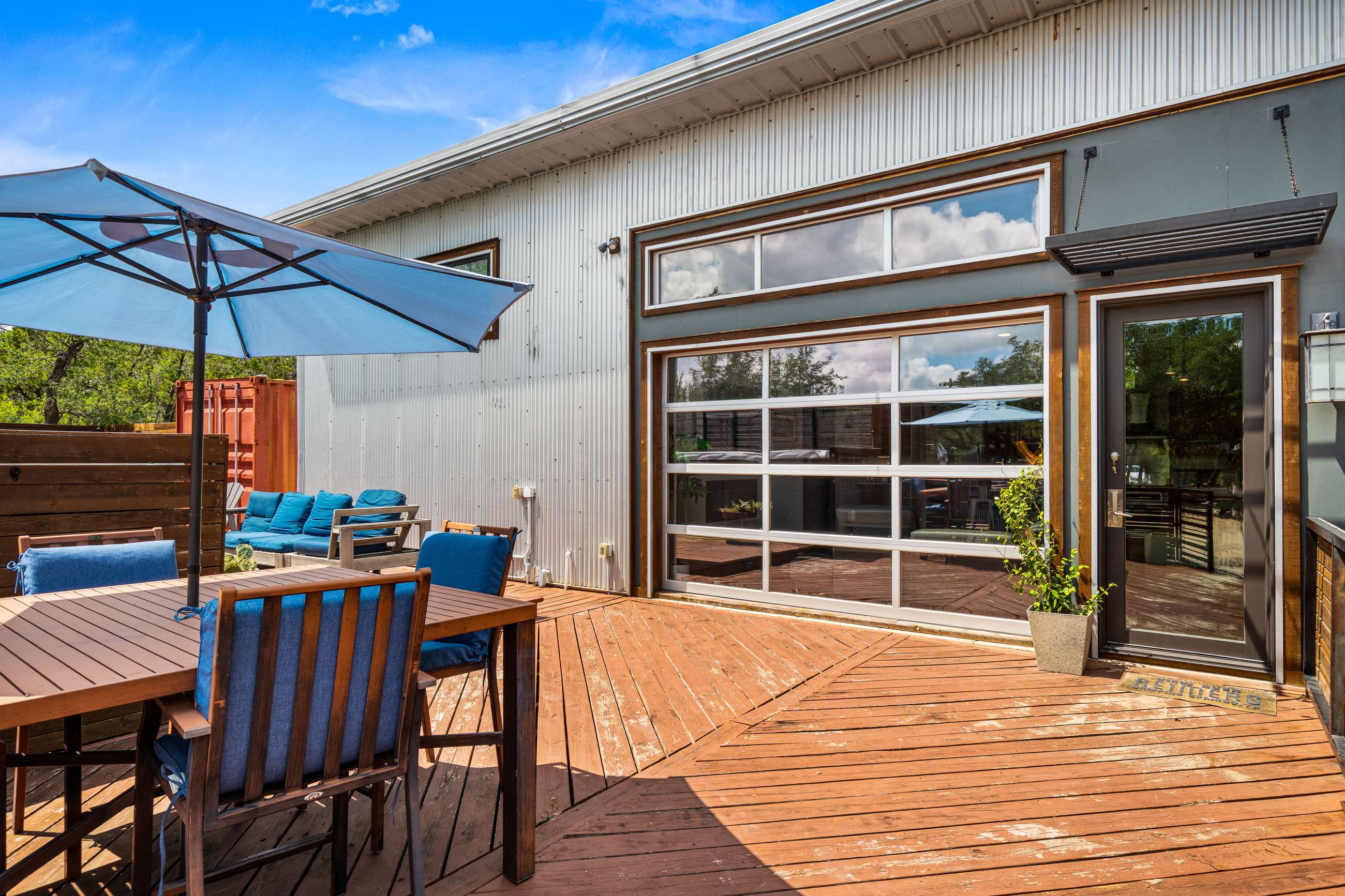 Wooden deck with an outdoor dining table and chairs under a blue umbrella, next to the modern house exterior featuring corrugated metal siding and a large glass garage-style door. Lush greenery and trees are visible in the background.
