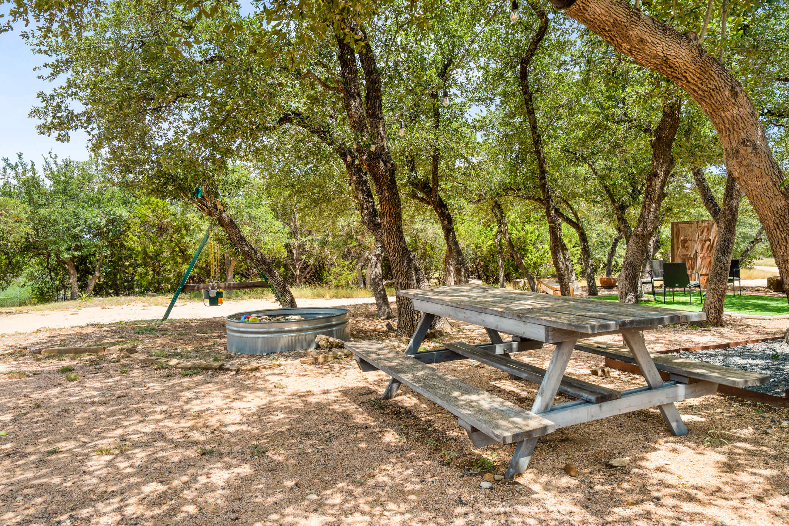 A shaded outdoor area with gravel ground features a wooden picnic table and a large metal stock tank, surrounded by numerous mature trees creating dappled sunlight.