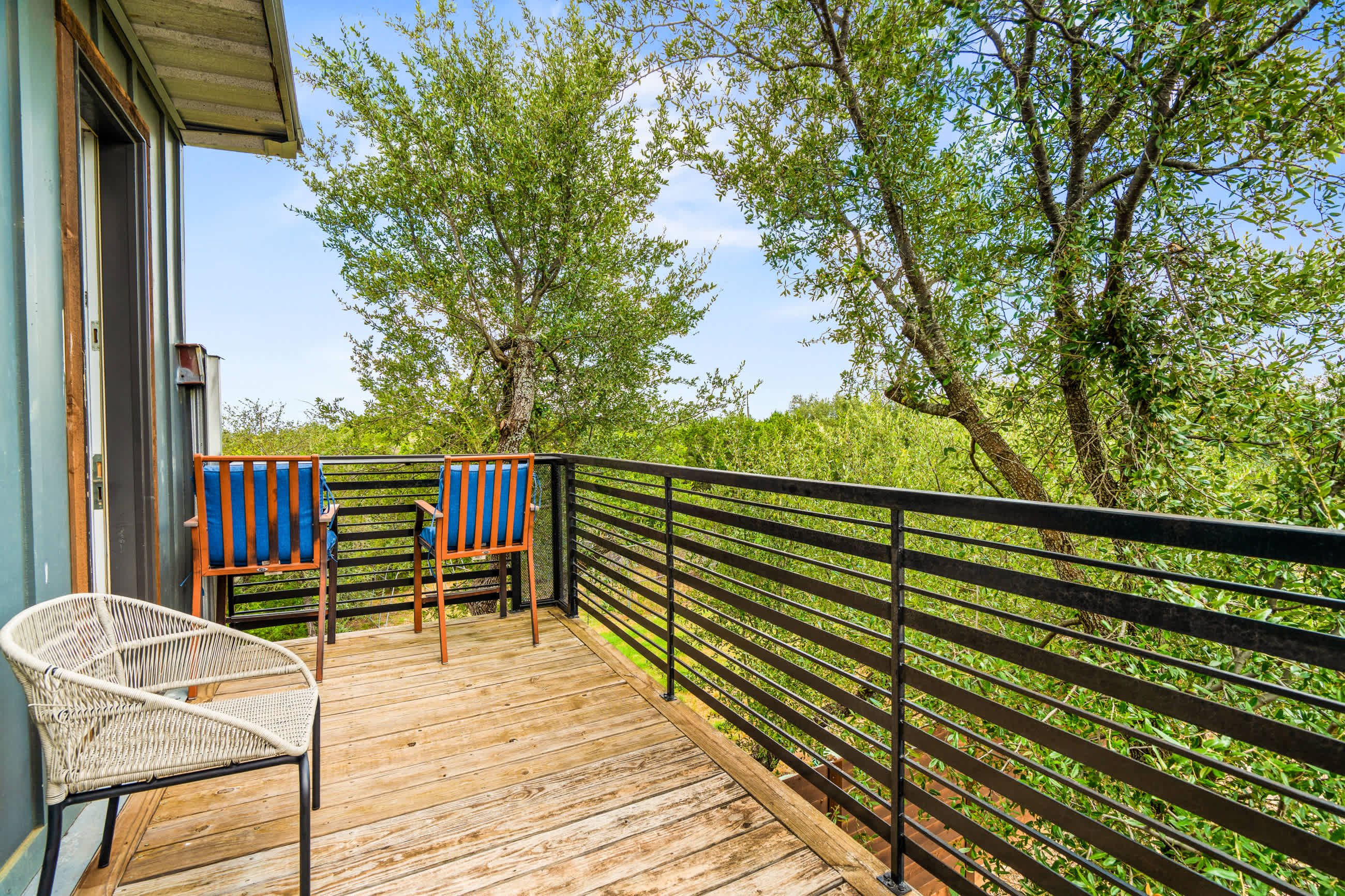 Small private balcony with a wooden floor and black metal railing, furnished with two chairs, offering a serene view of lush green trees and natural landscape under a bright blue sky.