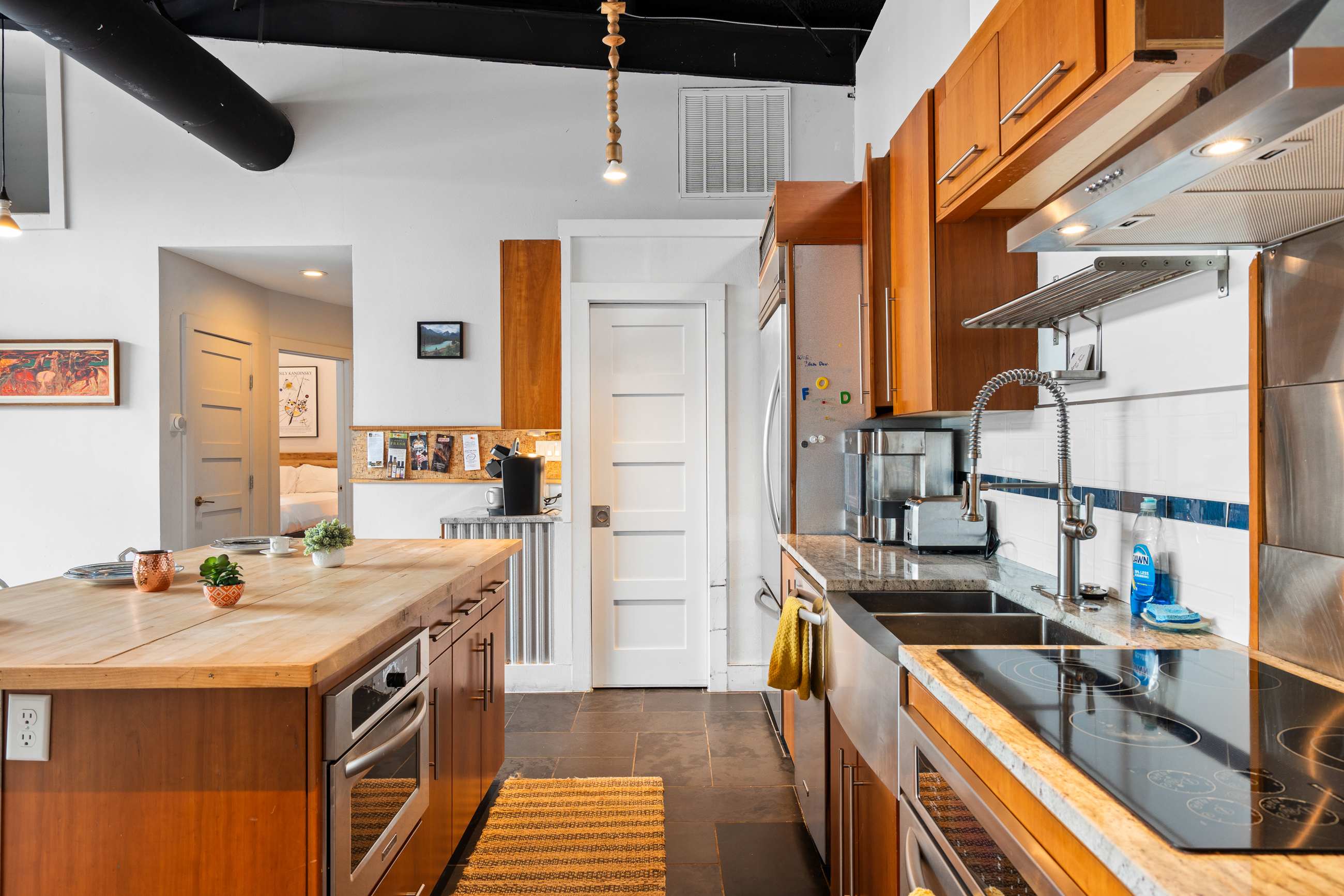 A modern kitchen with wood cabinetry, stainless steel appliances, and a large farmhouse sink. The counter space includes a cooktop, and a wooden island with a built-in oven is visible in the foreground.