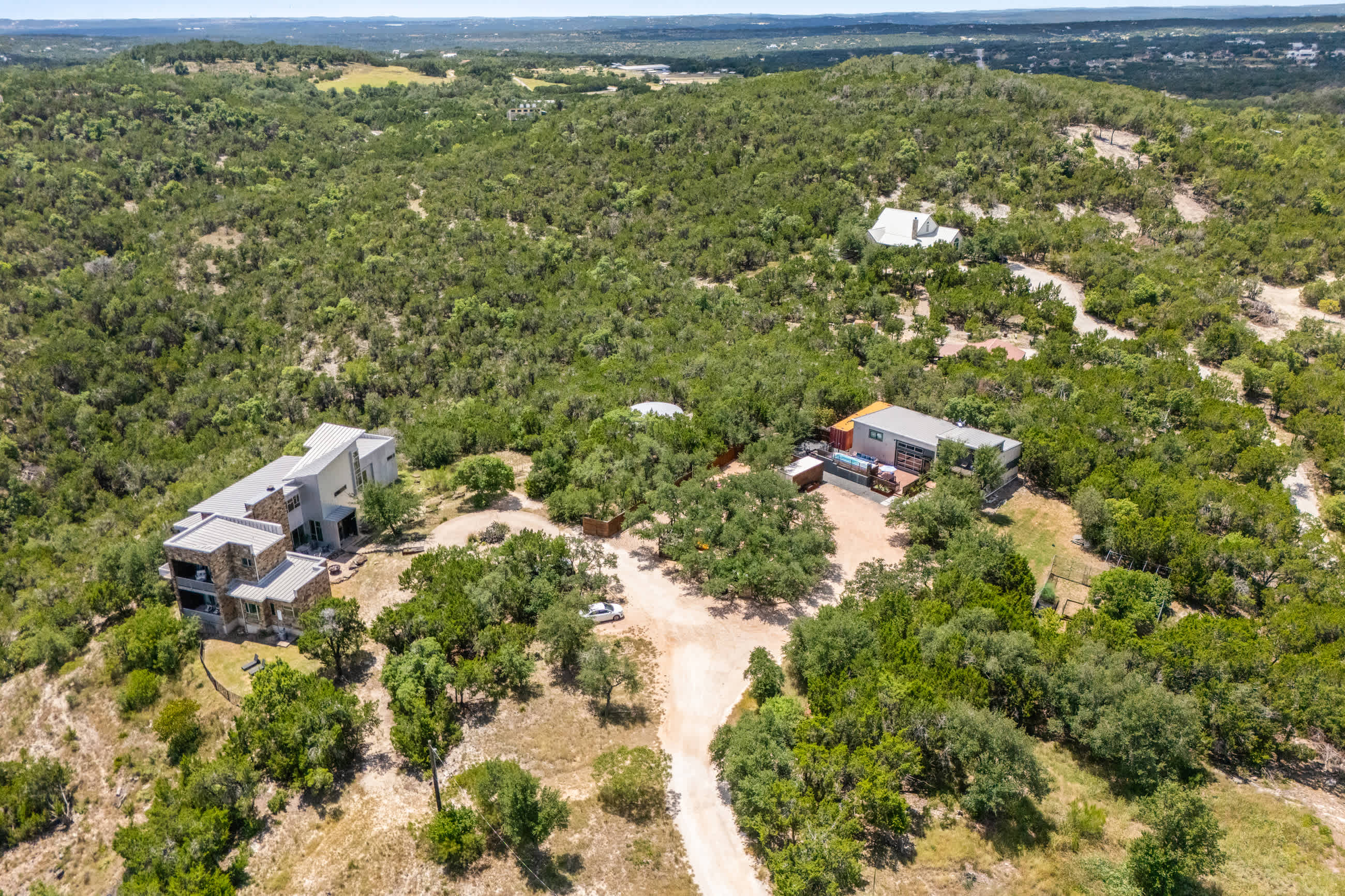 A wider aerial shot reveals two modern houses with grey roofs situated on a tree-covered hillside, connected by a dirt road, surrounded by vast natural landscape.