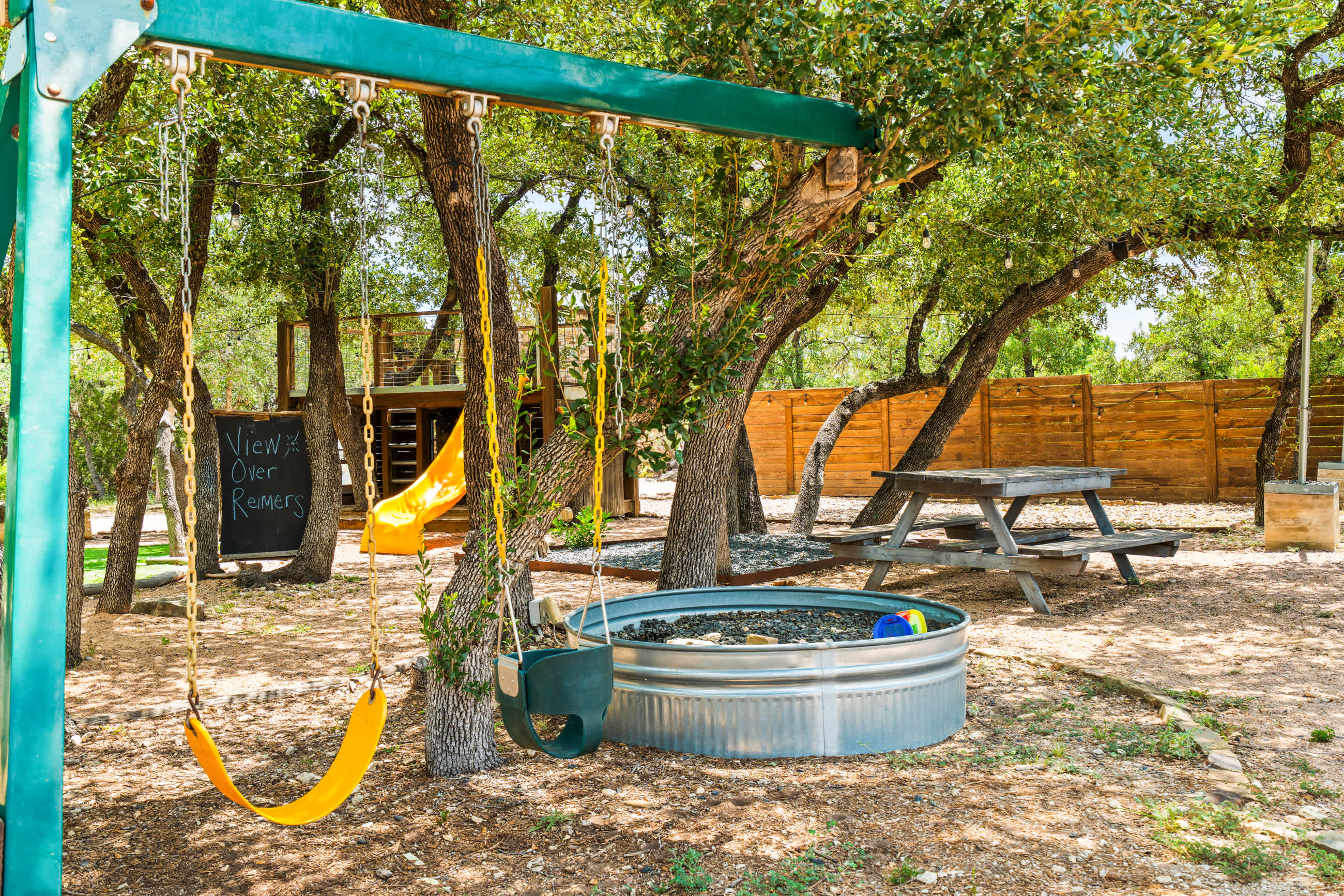 A children's play area under trees features a green swing set with yellow swings, a metal stock tank, and a wooden picnic table in the background, all on gravel ground.