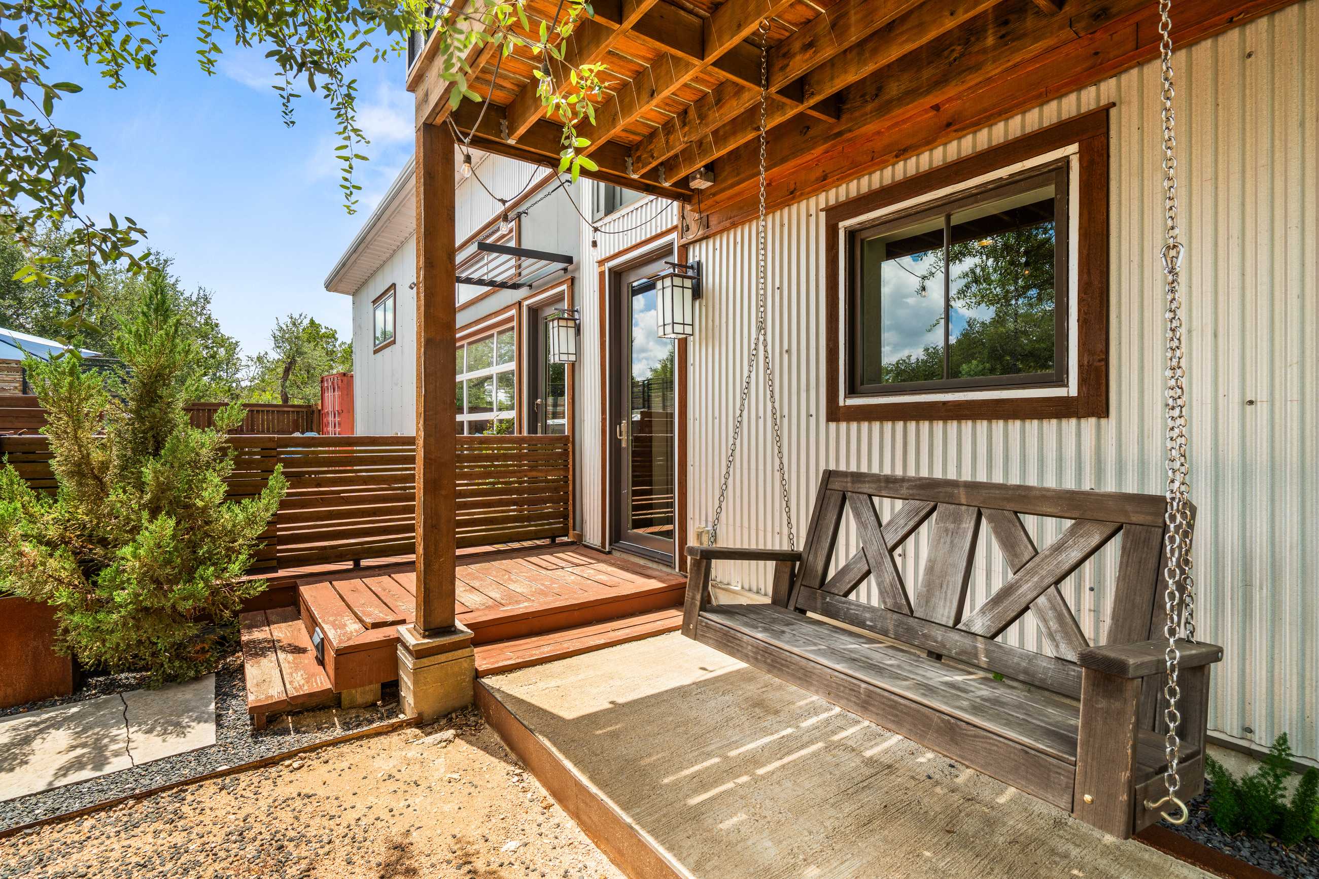 Covered porch area with a rustic wooden swing, adjacent to the house exterior with corrugated metal siding and a glass door. The area features a mix of gravel and concrete, with greenery providing a natural touch.