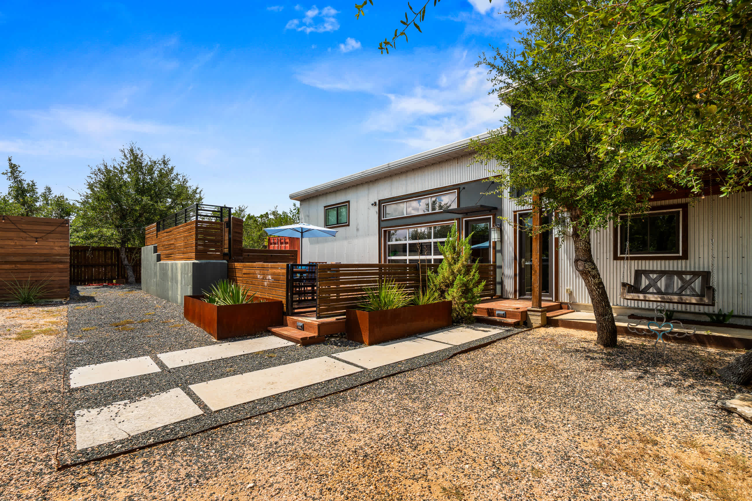 Front exterior of a contemporary house featuring corrugated metal siding and a prominent glass garage-style door, with a gravel driveway and concrete stepping stones leading to the entrance. Mature trees and modern landscaping with rusted metal planters enhance the curb appeal.
