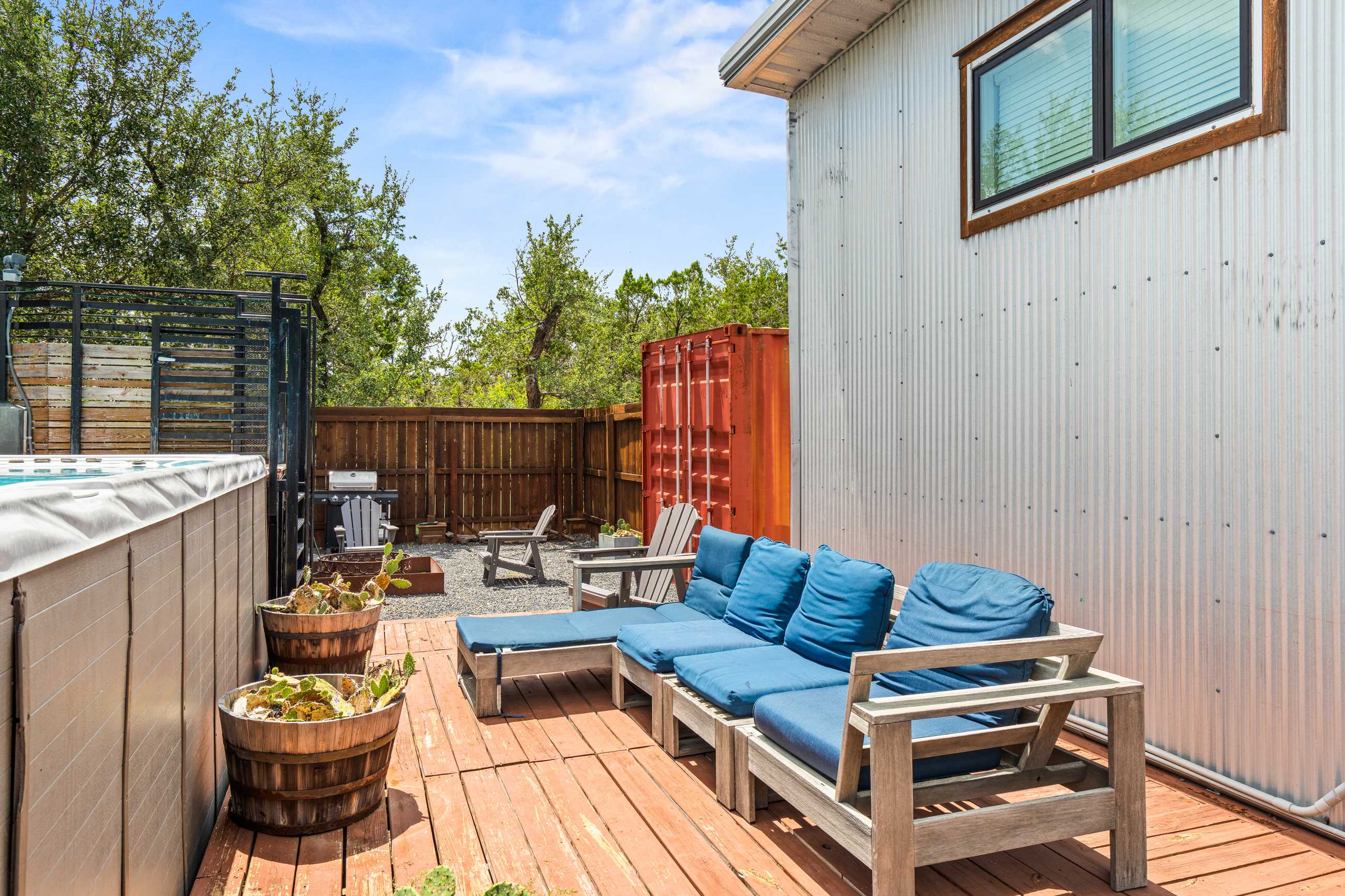 Wooden deck area with blue cushioned lounge seating next to a swim spa, with a corrugated metal wall of the house and a wooden fence in the background.