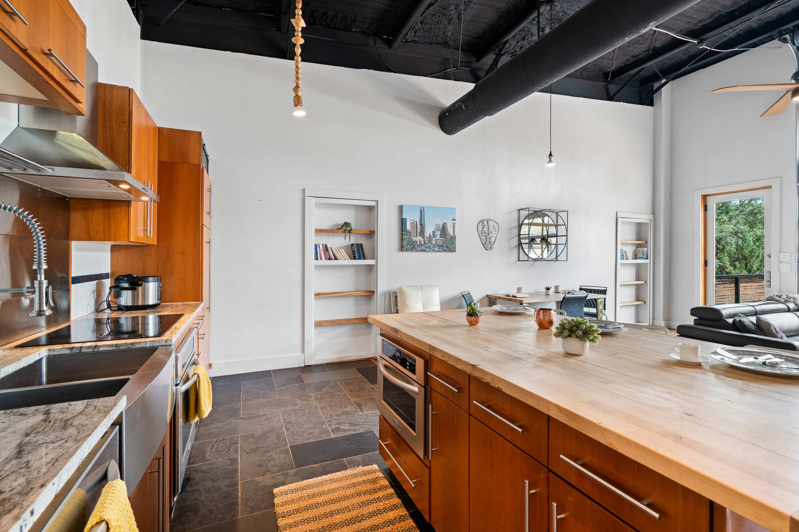 A wide view of the open-concept kitchen and dining area featuring a large wooden island with an integrated microwave, dark tile flooring, and white walls with exposed black ceiling ducts, leading to a dining table and built-in shelves.