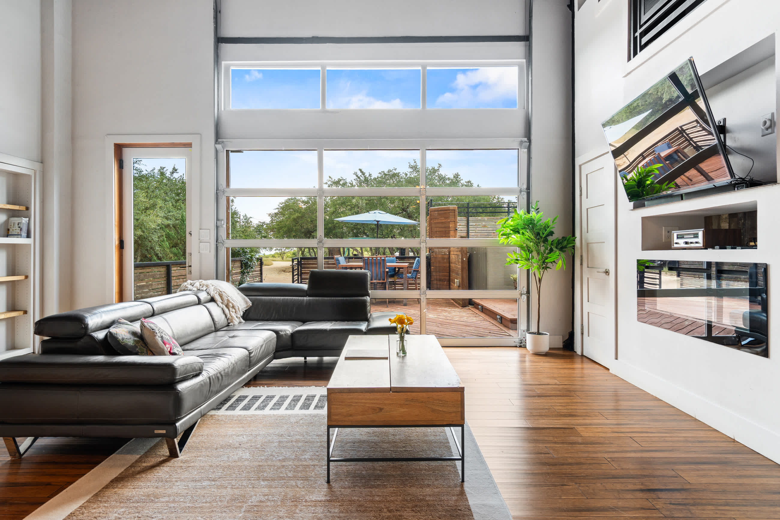 A modern living room with a dark leather sectional sofa and a wooden coffee table, facing a large glass garage-style door that opens to an outdoor deck with seating and trees. The room features dark wood floors and a wall-mounted TV.