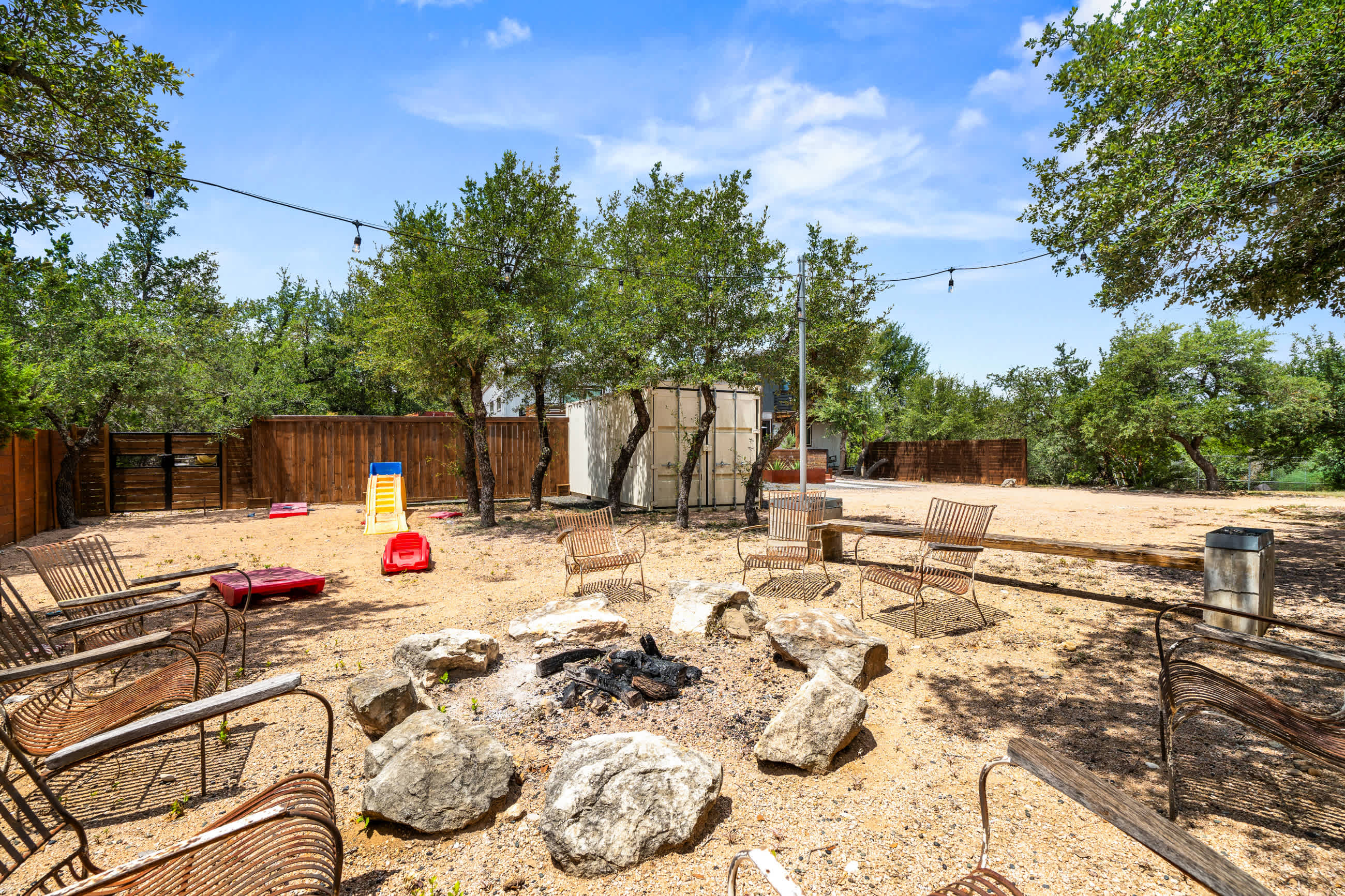 A sandy outdoor area features a large rock-ringed fire pit surrounded by several rustic metal chairs, with a wooden fence and trees in the background under a clear blue sky.