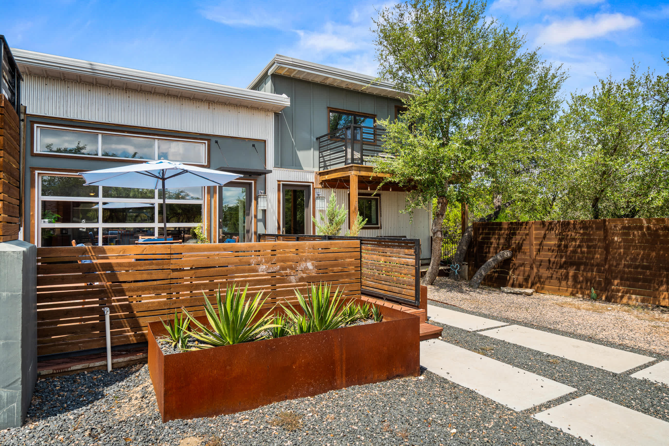 Exterior view of the house showcasing modern architecture with corrugated metal siding, a small upper balcony, and a landscaped entrance with wooden slatted fencing and rusted metal planters. A gravel path with concrete stepping stones leads to the front door.
