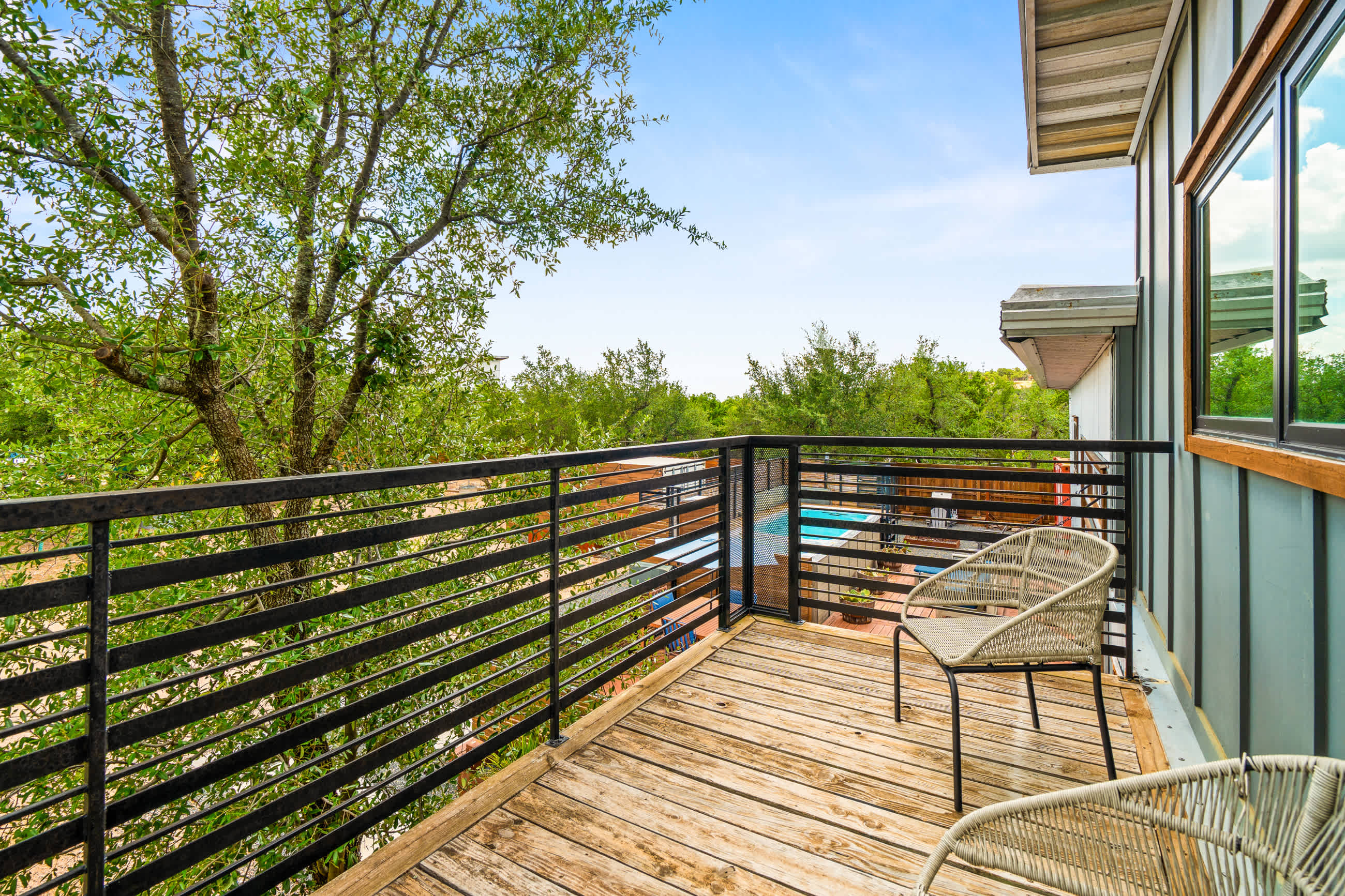 Outdoor balcony with weathered wooden planks and a black metal railing, offering views of lush green trees and a distant swimming pool area. Two light-colored woven chairs are placed on the balcony.