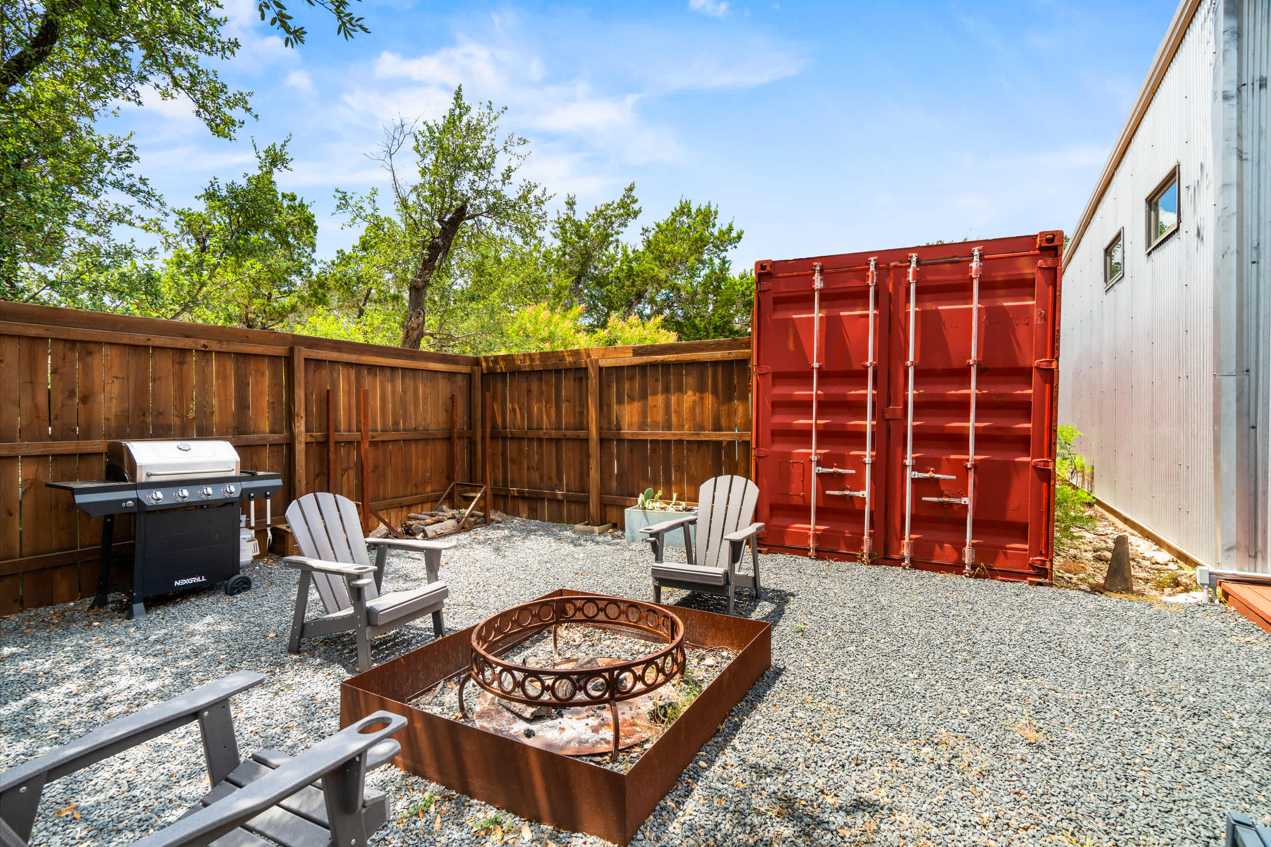 Gravel outdoor area featuring a large metal fire pit, gray Adirondack chairs, a BBQ grill, and a red shipping container against a wooden privacy fence.