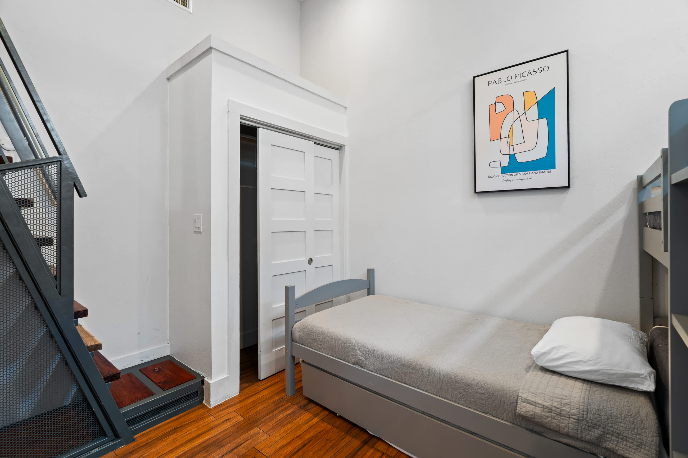 View of a kids' bedroom with a gray single bed and a trundle, dark hardwood floors, and white walls. A closet with white sliding doors is visible next to an industrial-style staircase.