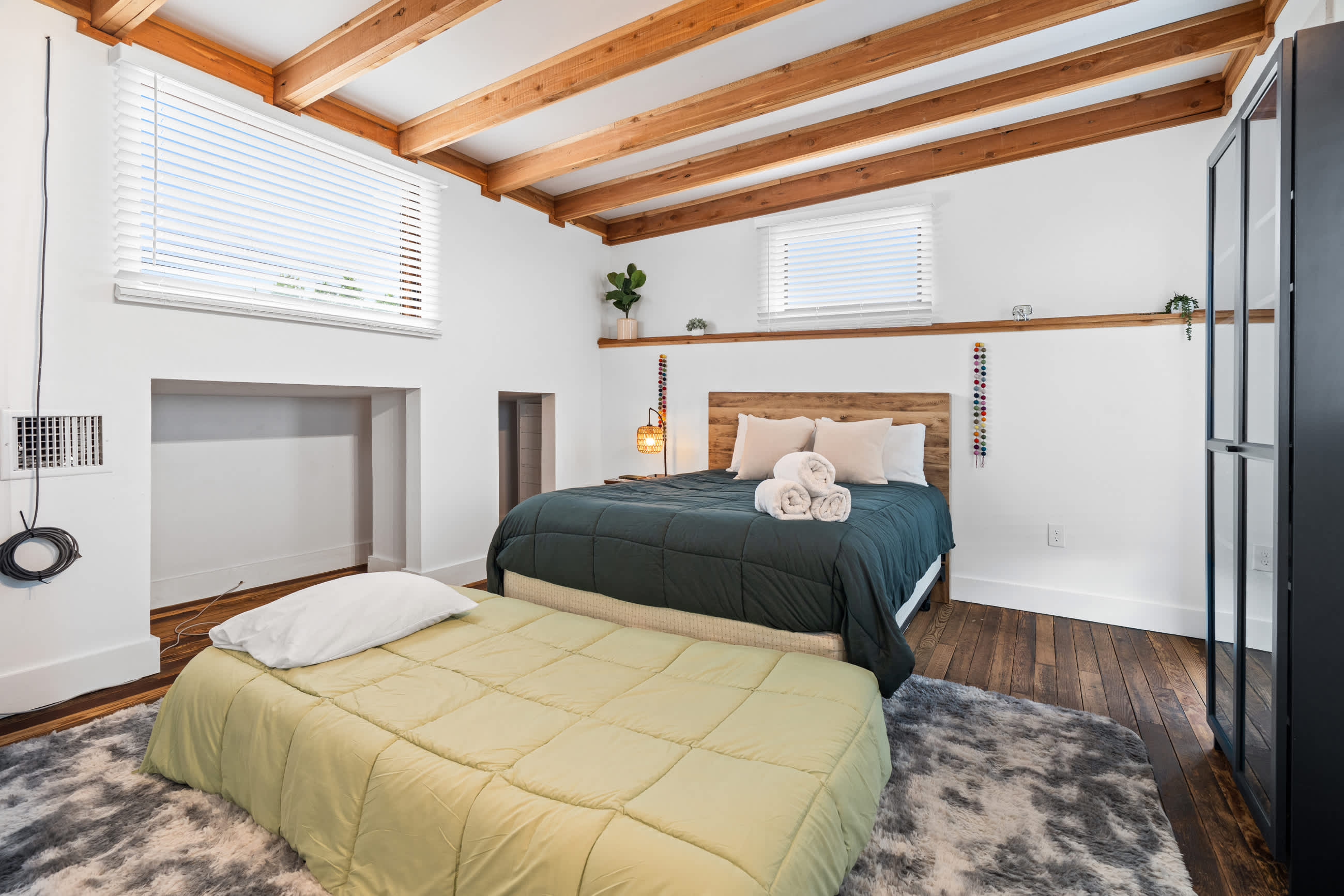 A full view of the bedroom with a queen bed, a light green blanket, exposed wooden ceiling beams, and two windows, featuring a small shelf above the bed and dark wood flooring with a grey rug.