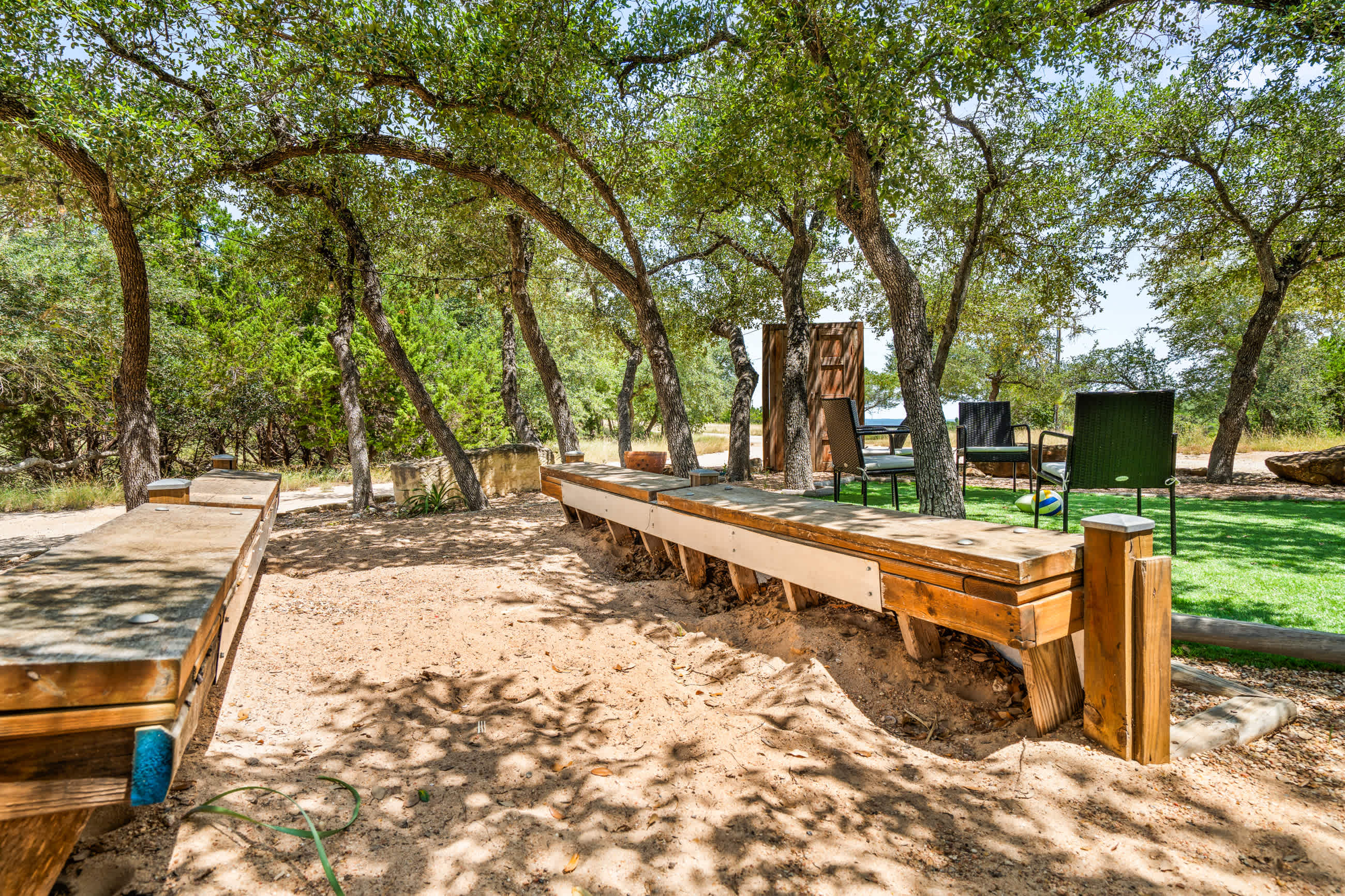 A long wooden bench with a built-in planter runs along a gravel path, shaded by numerous trees, leading towards a grassy area with outdoor seating.