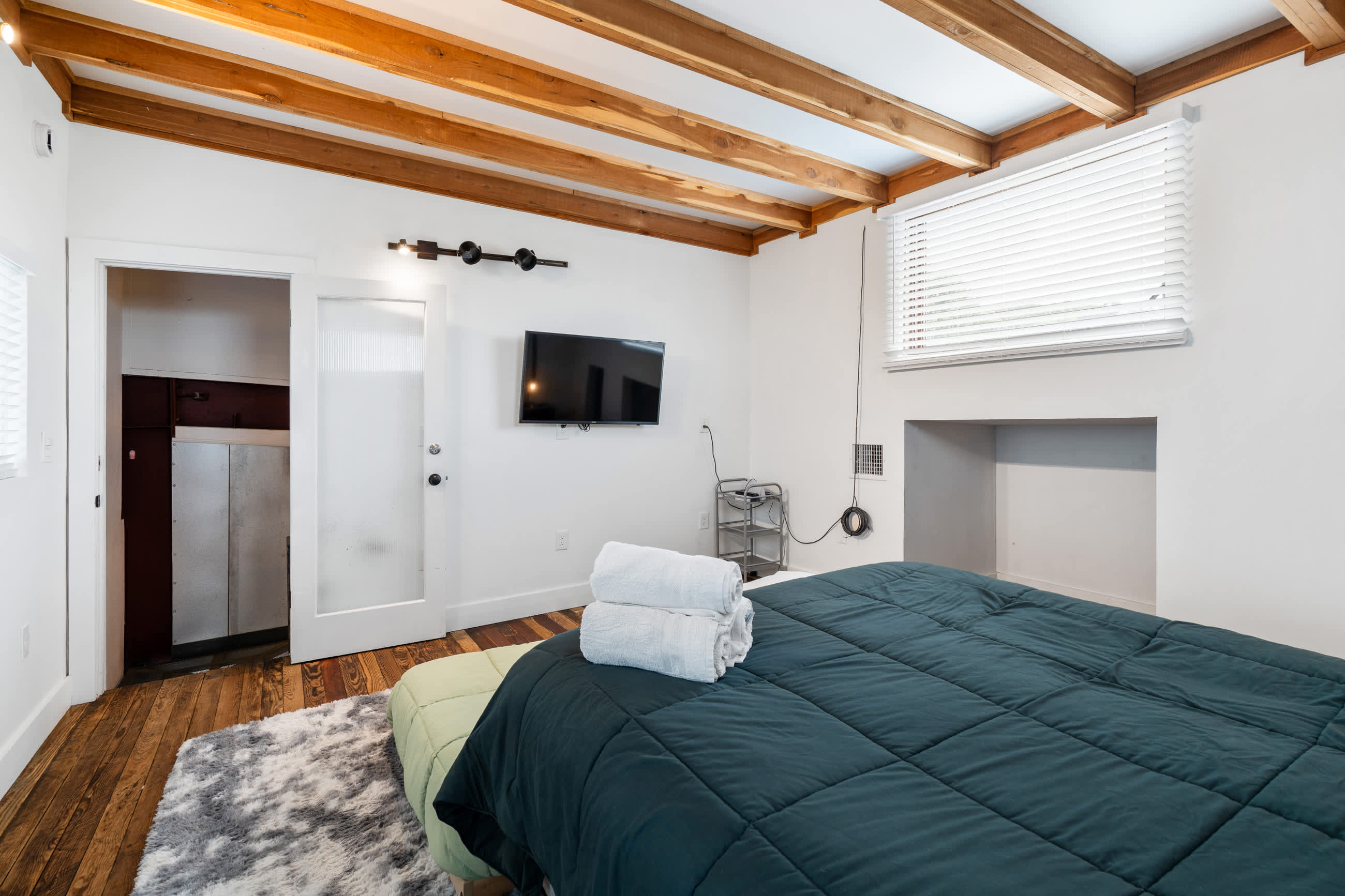 Cozy bedroom featuring a dark teal comforter on a bed, a white shaggy rug, and exposed wooden ceiling beams. A wall-mounted TV is visible, along with a window with white blinds and a partial view into an adjacent room.