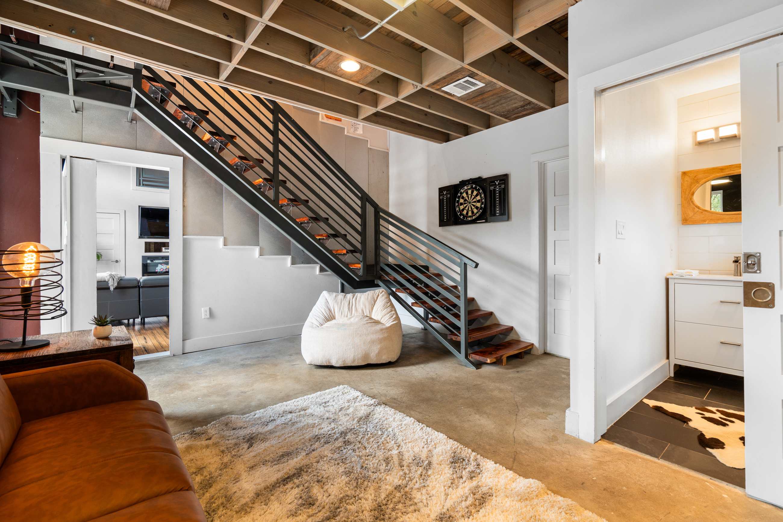 An industrial-style staircase with metal railings leads to an upper level, adjacent to a dartboard on the wall and a bathroom entrance. The area features concrete floors and an exposed beam ceiling.