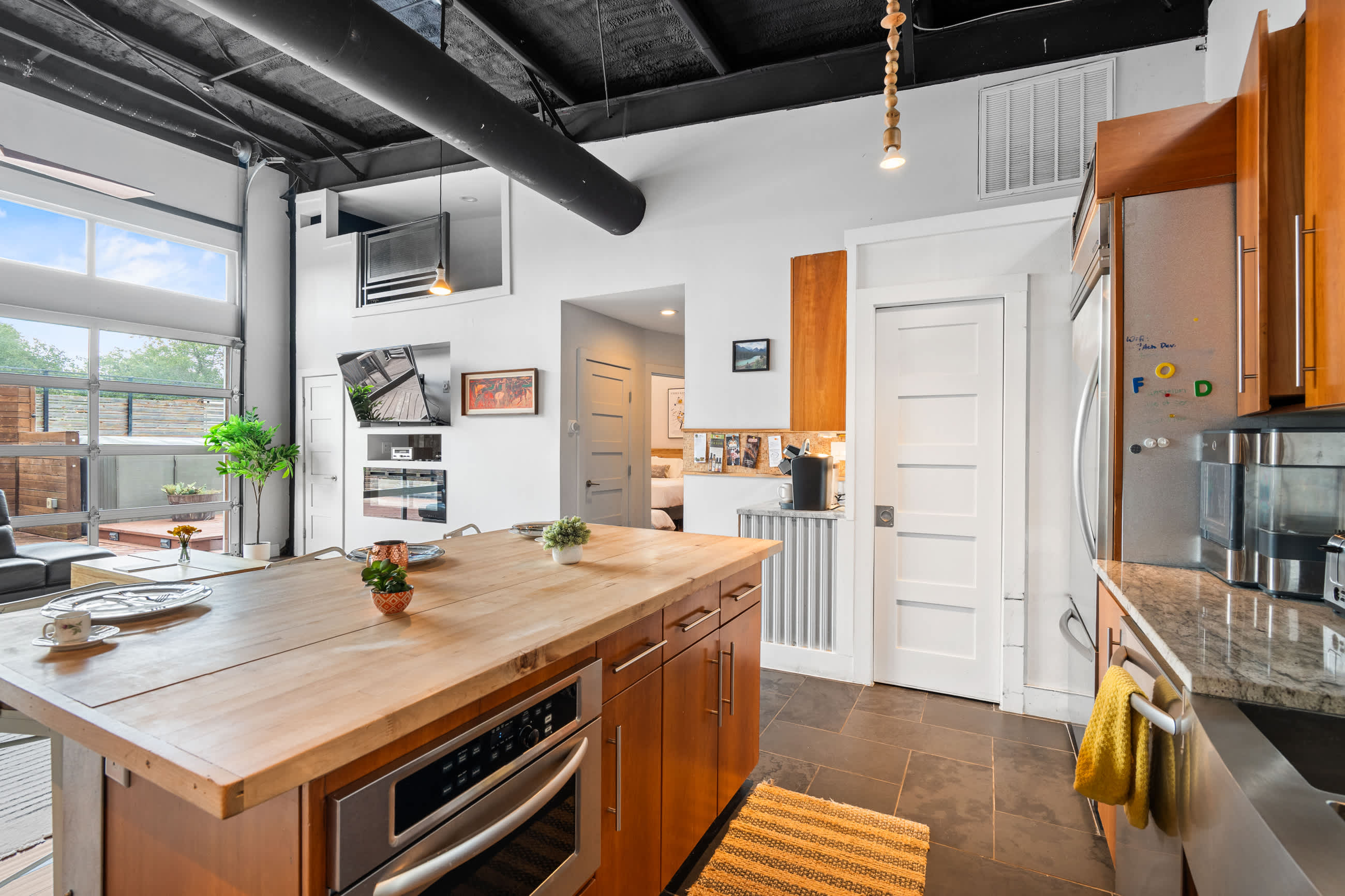 A view of the kitchen from the dining area, showing the stainless steel refrigerator, light wood cabinetry, and a coffee station, with a white door leading to another room and a large window above the kitchen area.