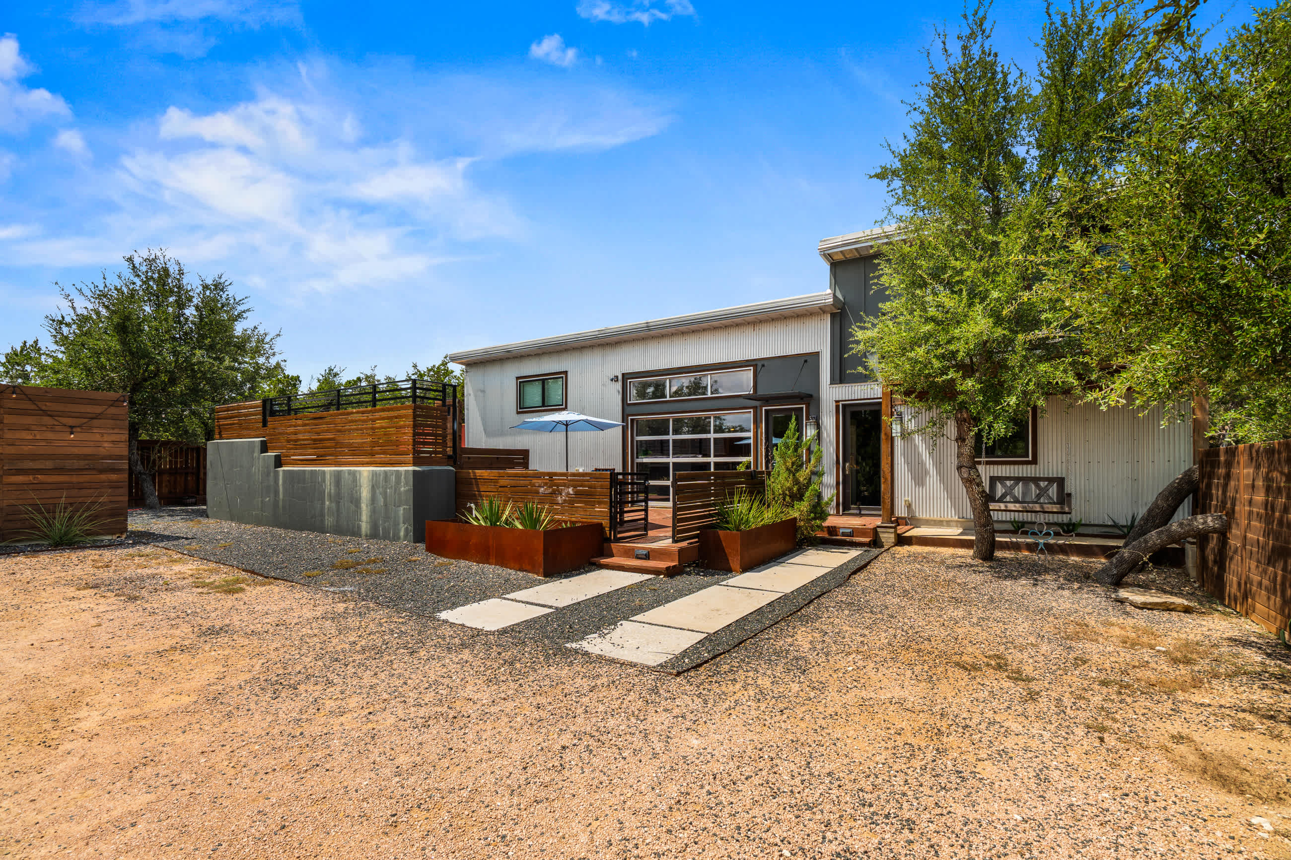 Front exterior of a modern house with corrugated metal siding and a large glass garage-style door, set against a gravel driveway. Landscaped with concrete stepping stones, dark gravel, and rusted metal planters with greenery, under a bright blue sky.