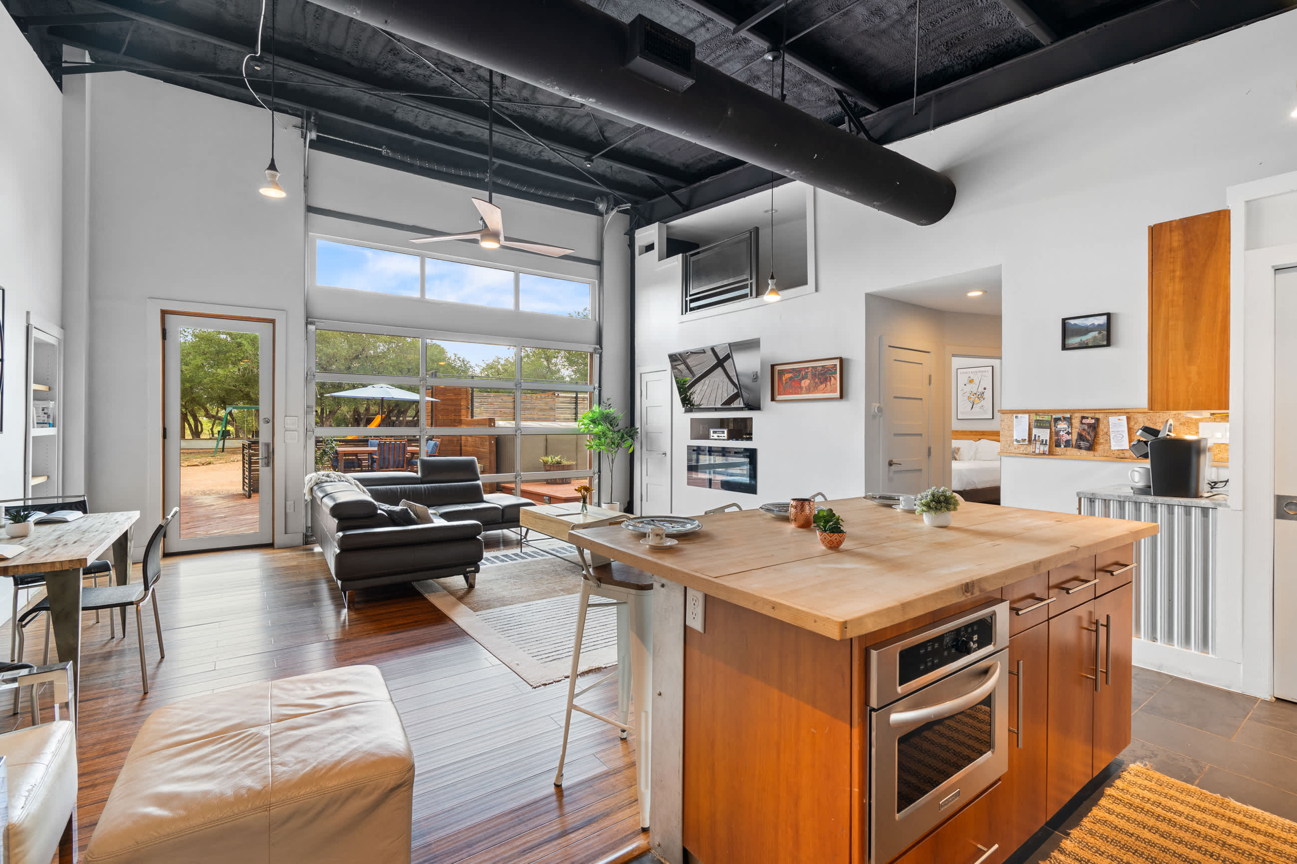 A bright, open-concept living space featuring a modern kitchen with a large wooden island and bar stools, flowing into a living area with a dark sectional sofa. A large glass garage-style door provides abundant natural light and access to the outdoors.