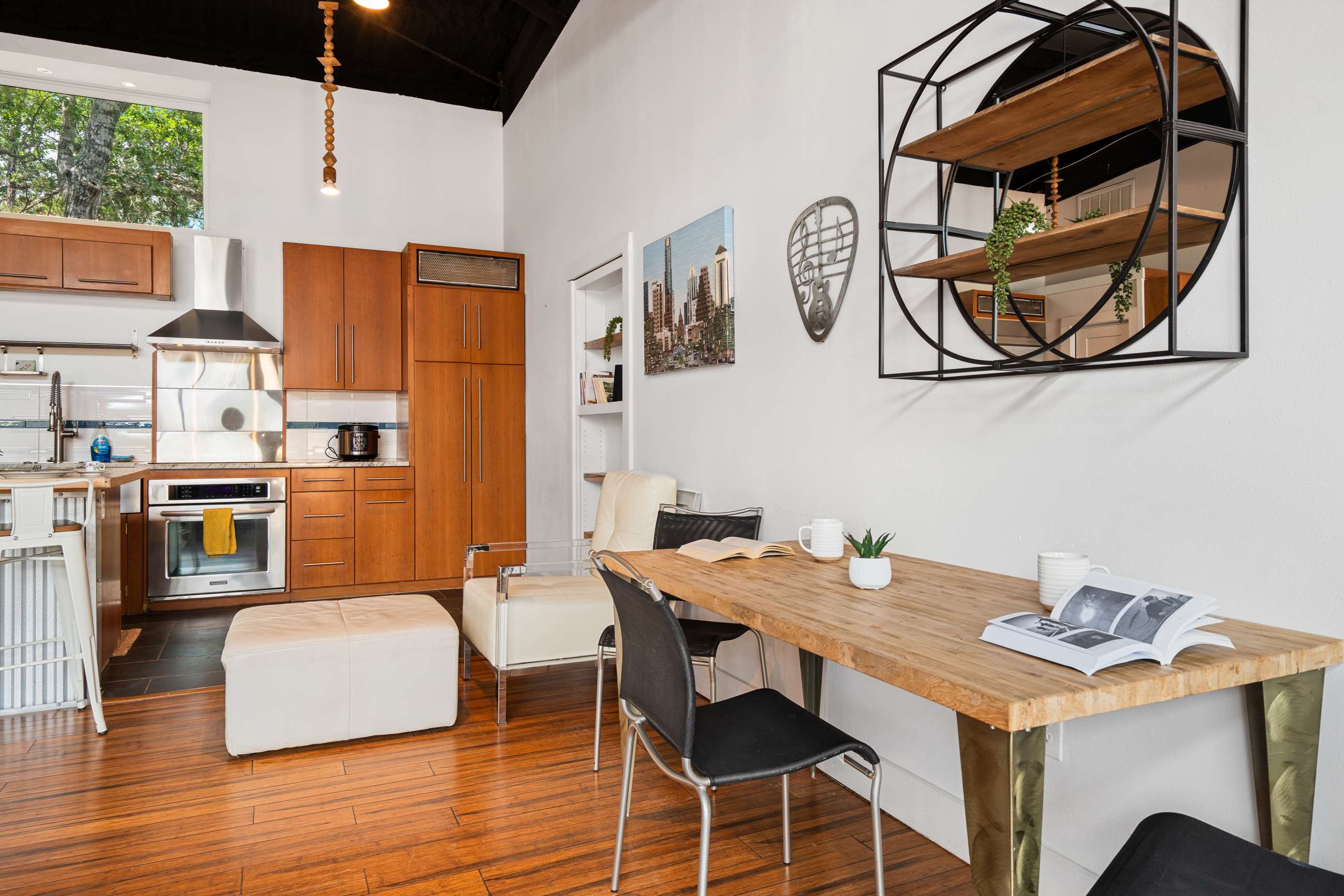 An open-plan kitchen and dining area with a wooden dining table and black chairs, adjacent to a modern kitchen featuring wood cabinets, stainless steel appliances, and a central island with white stools. The space has dark wood floors and white walls.