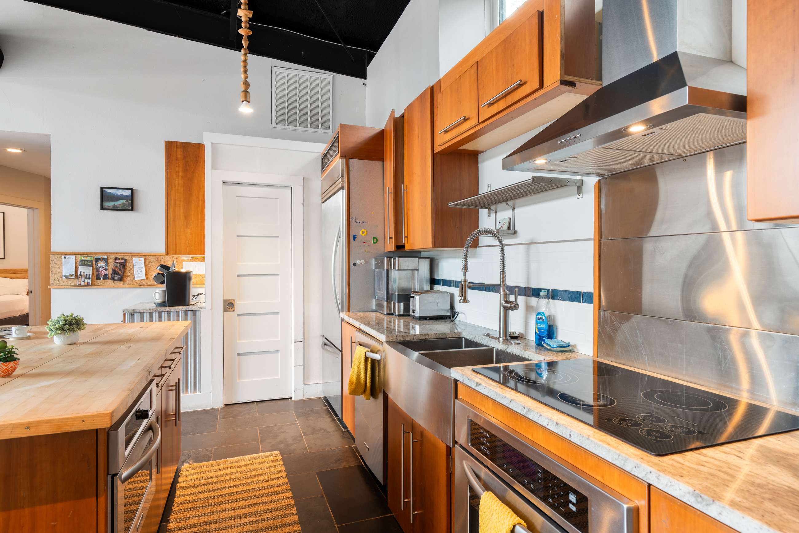 A close-up of the kitchen area showcasing the stainless steel refrigerator, double sink, electric cooktop, and built-in oven, with light wood cabinets and a white subway tile backsplash, and dark tile flooring.