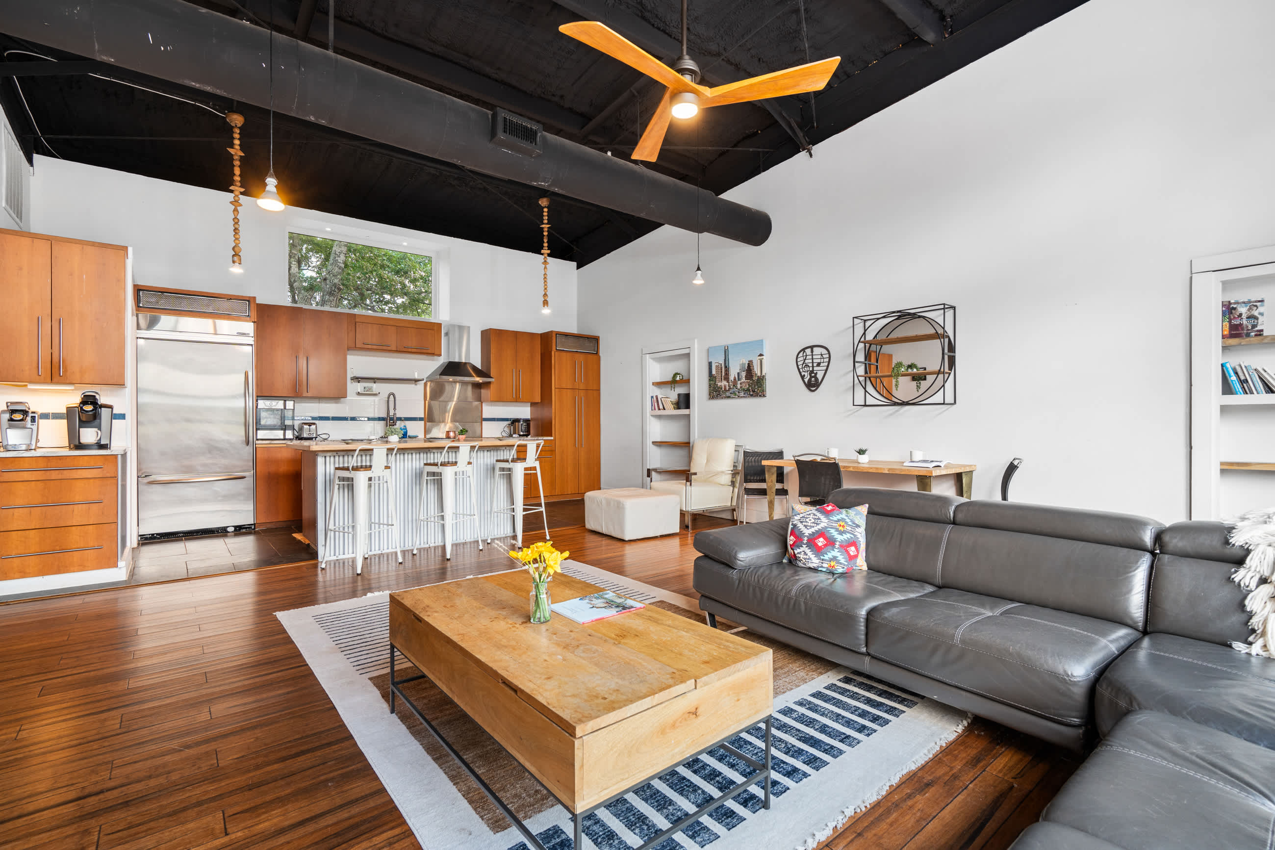 An open-concept living area features a gray sectional sofa, a light wood coffee table, and a modern kitchen with wood cabinets and stainless steel appliances. The space has dark wood floors, white walls, and a black exposed ceiling with a ceiling fan.