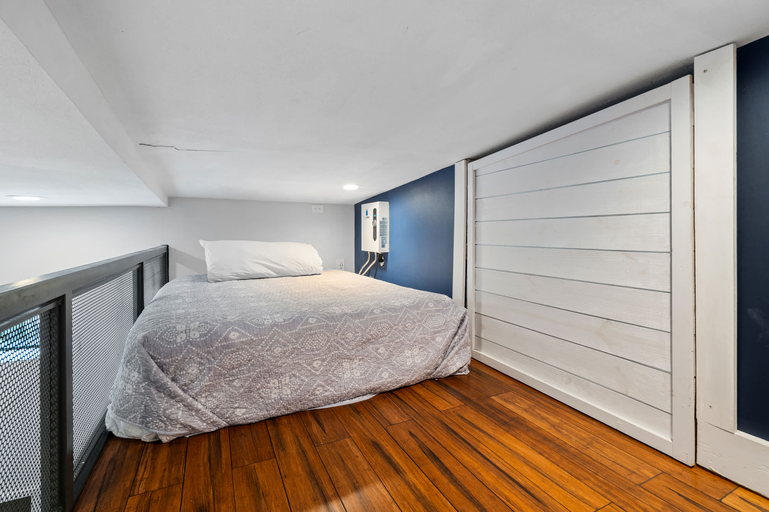 Loft bedroom with a bed on dark hardwood floors, featuring a white shiplap accent wall and a dark blue wall. A water heater is mounted on the blue wall, and a metal mesh railing separates the loft from the lower level.