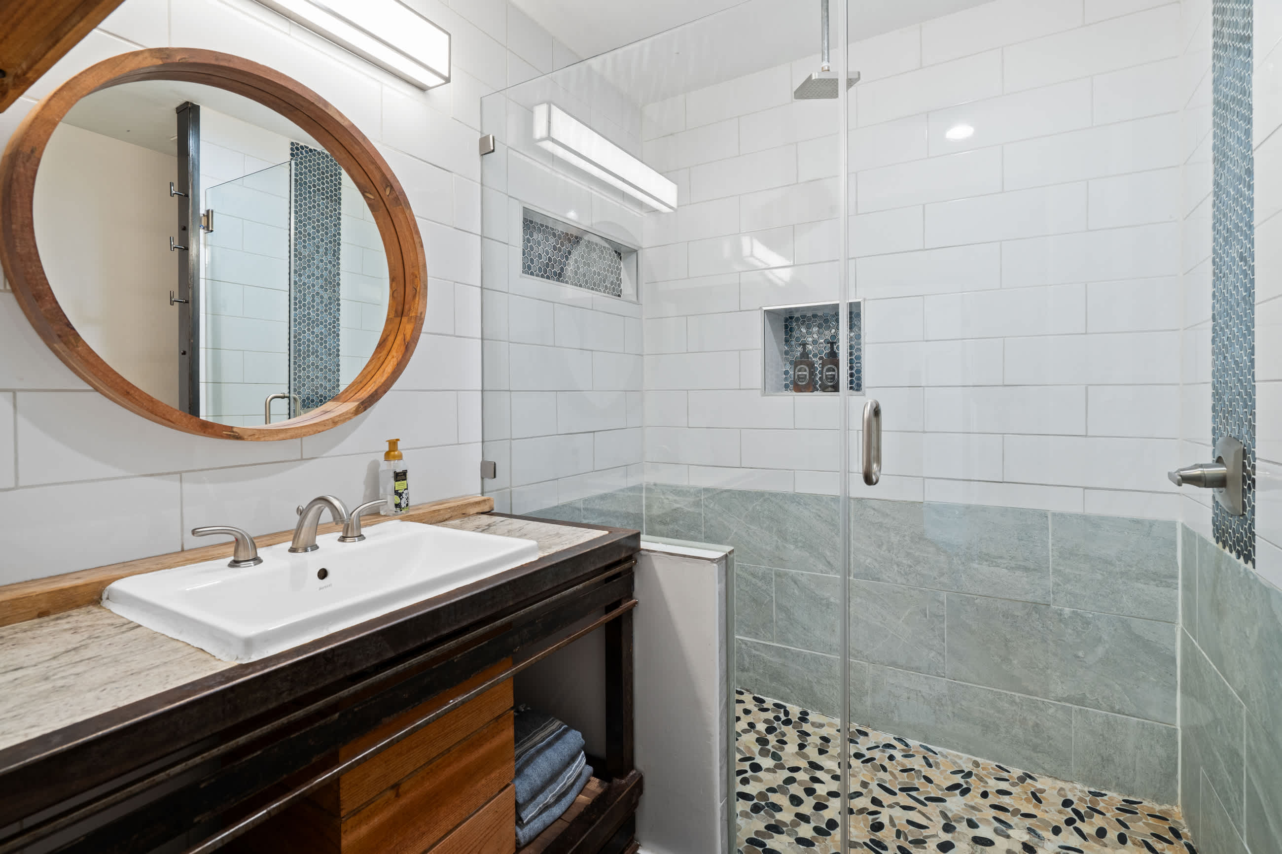 A closer view of the bathroom vanity and walk-in shower, highlighting the white subway tiles, pebble shower floor, and the dark wood vanity with open shelving for towels, next to a sliding barn door.