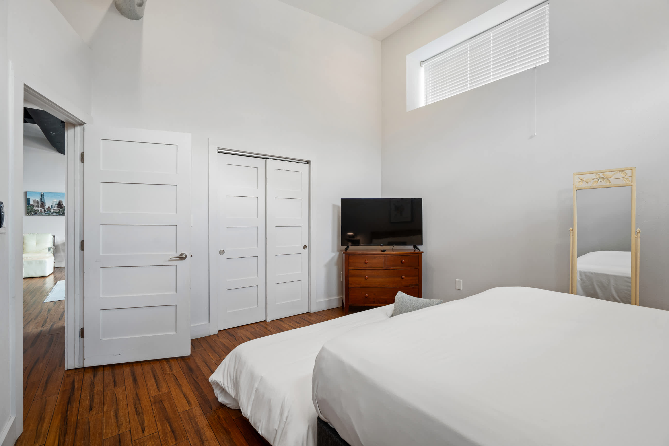 Bright bedroom with a large bed covered in white bedding, dark hardwood floors, and white walls. A wooden dresser with a TV sits opposite the bed, and a tall mirror leans against the wall.