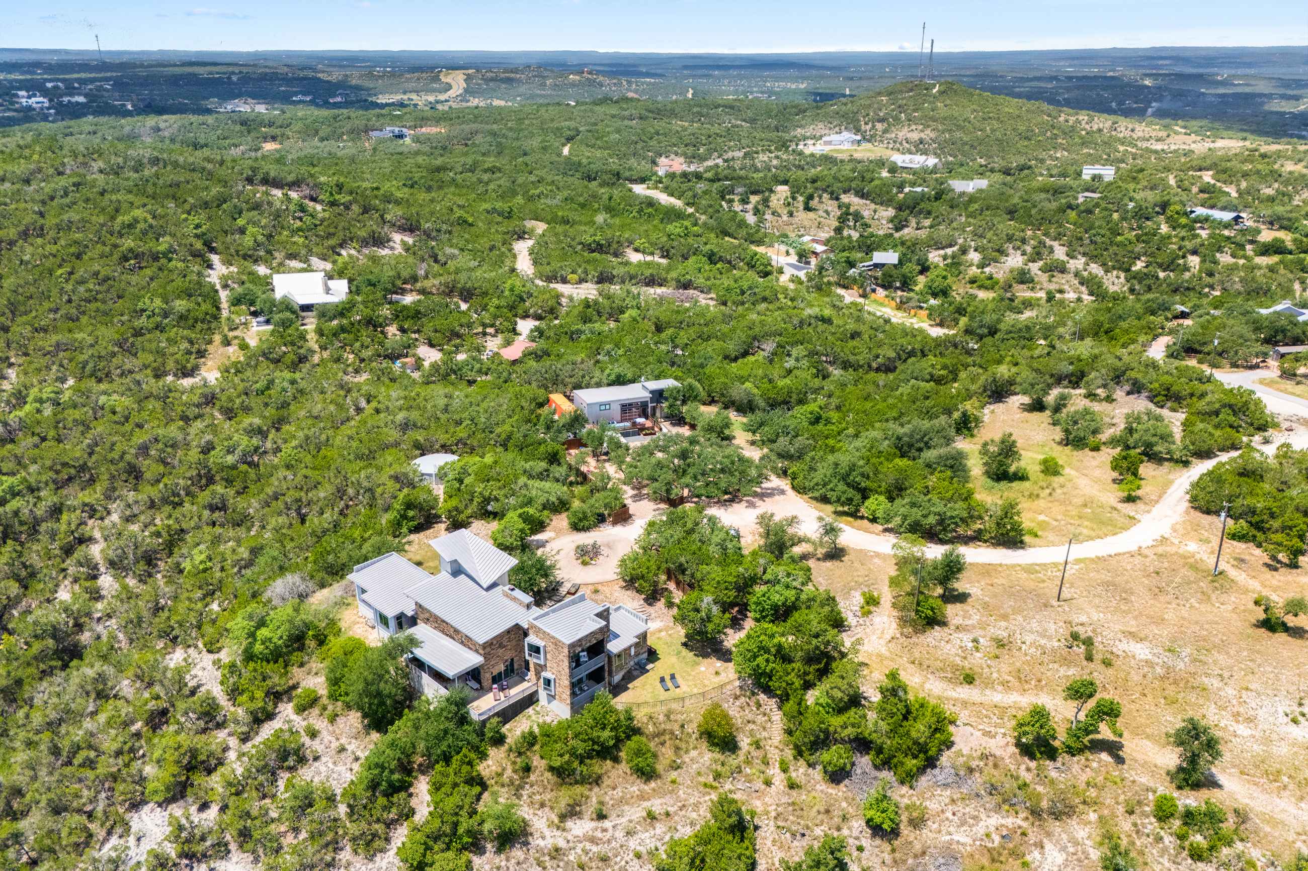 An aerial view showcases a modern house with a grey roof nestled among dense green trees on a hillside, with a dirt road leading up to the property.