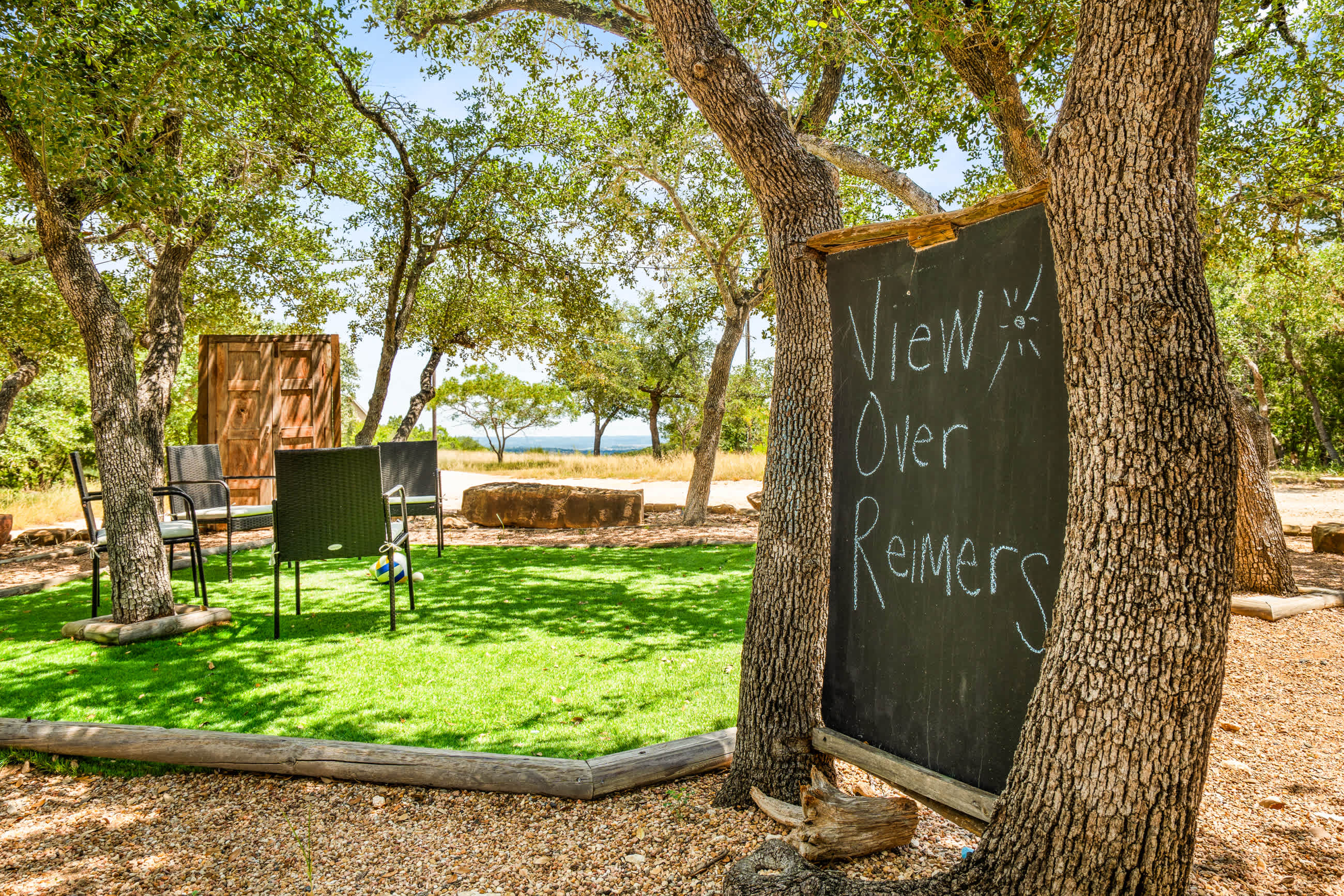 A chalkboard sign under a tree points towards a grassy area with black outdoor chairs and a distant view of a body of water, suggesting a scenic overlook.
