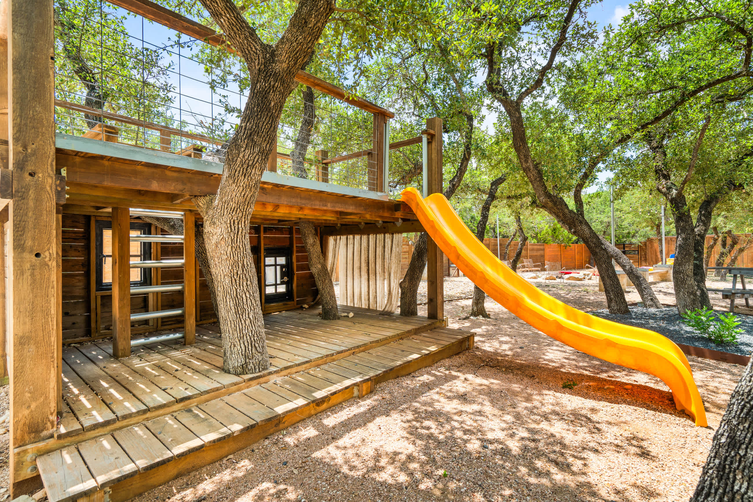 A wooden play structure with a bright orange slide is nestled among trees, featuring a raised platform with railings and a lower deck area, all on gravel ground.