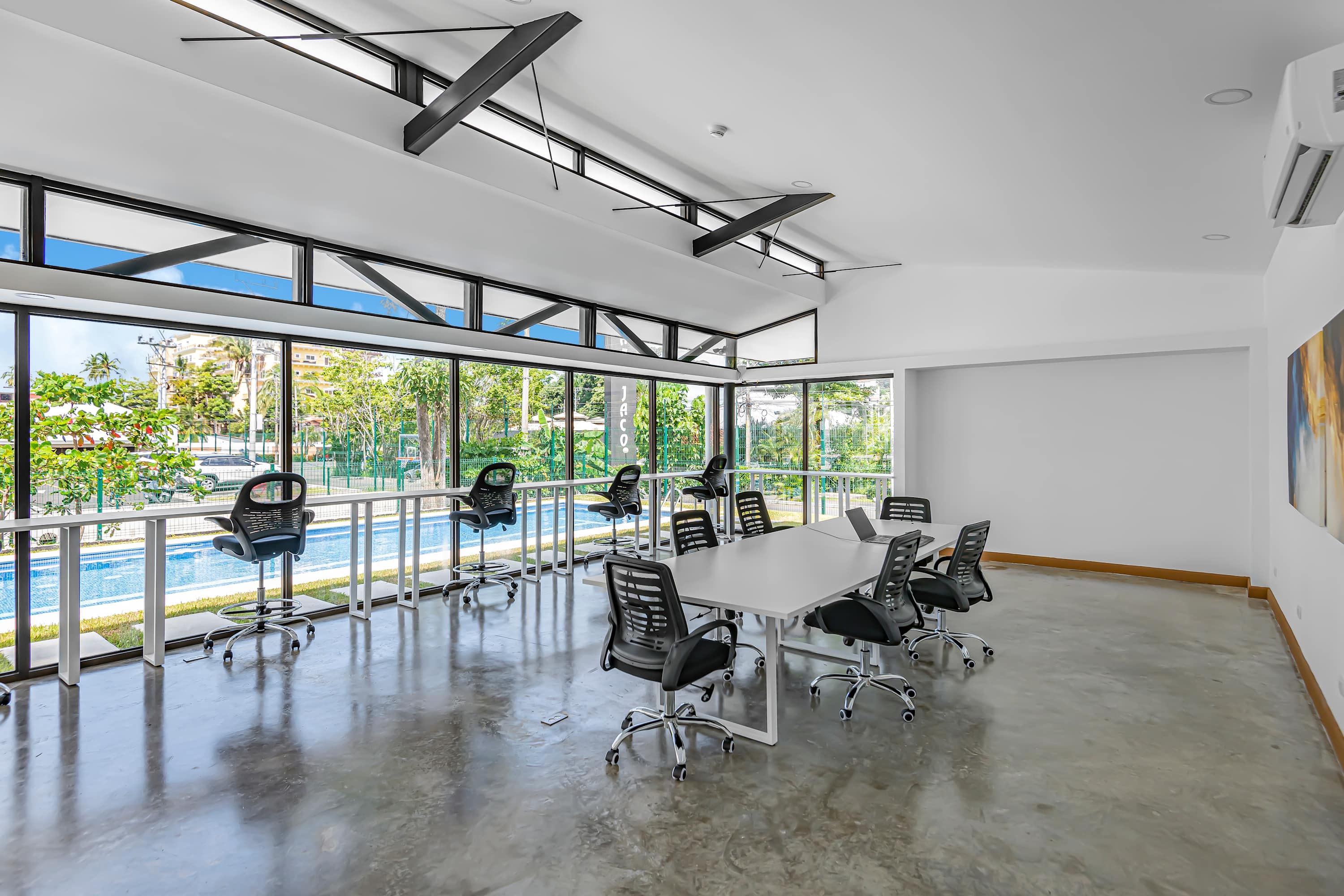 A bright, modern co-working space featuring a long white conference table with multiple black ergonomic chairs, set on a polished concrete floor. Large floor-to-ceiling windows offer views of the outdoor pool and tropical surroundings.