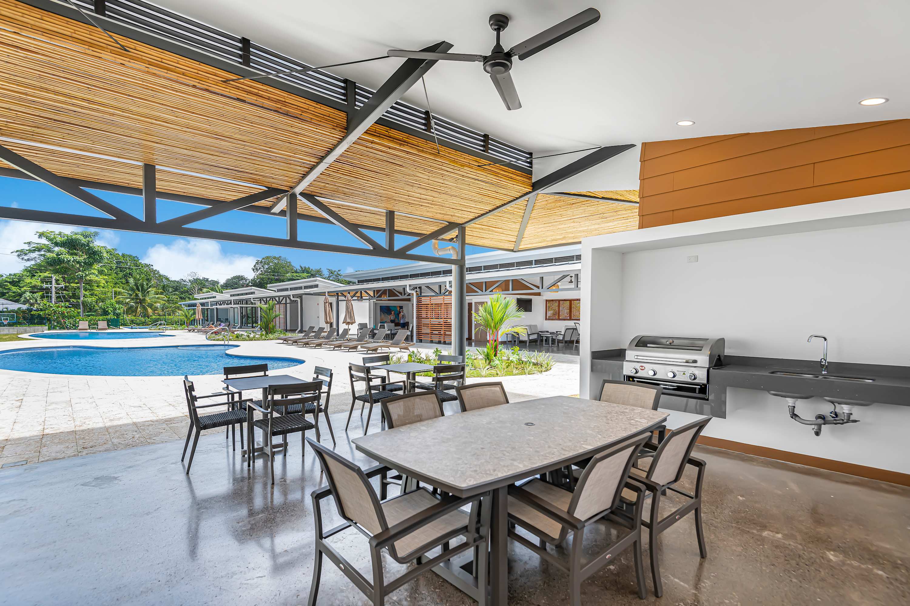 Covered outdoor dining and BBQ area with a large table and chairs, a gas grill, and a sink, overlooking the communal pool area. The ceiling features natural wood slats.