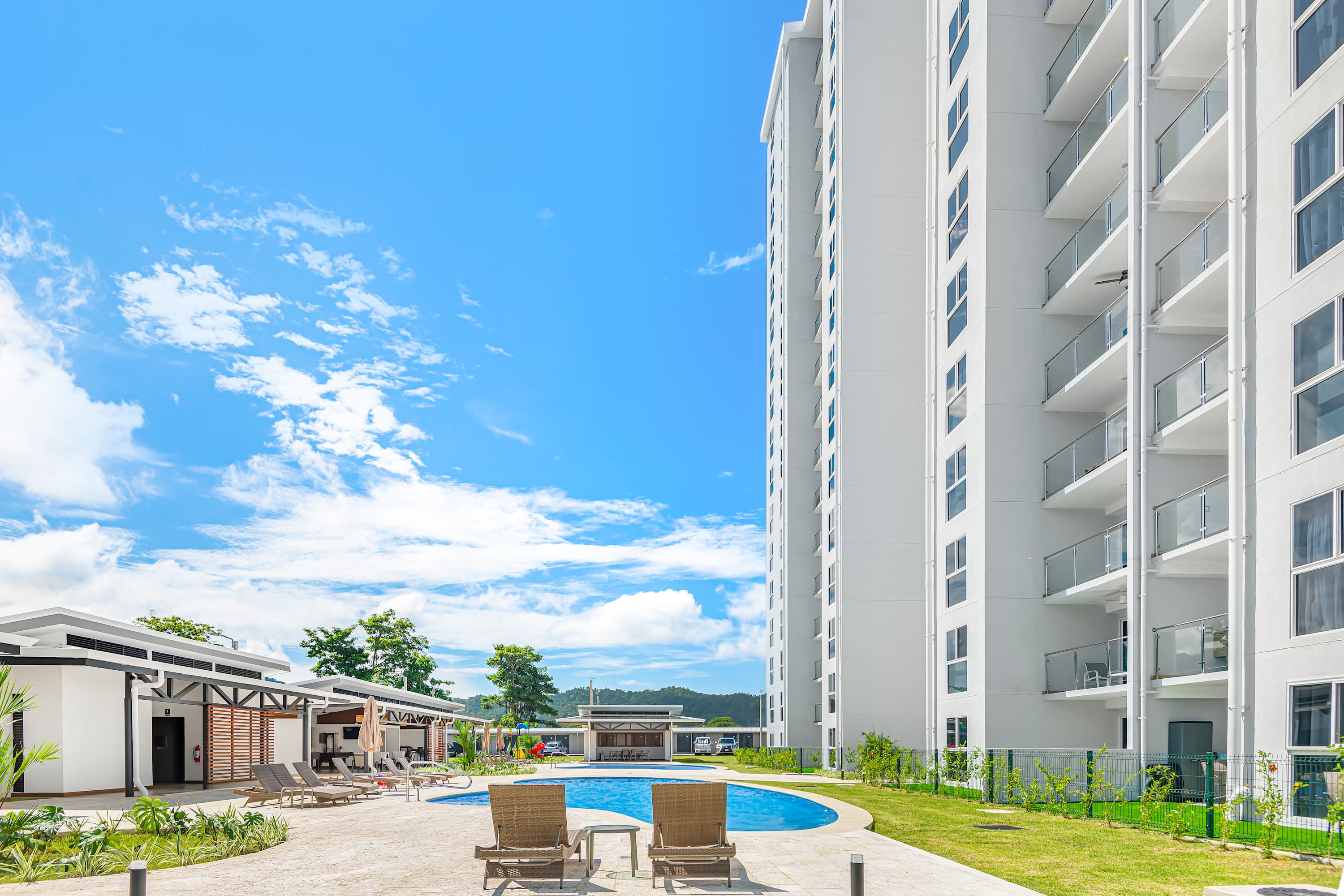 Expansive outdoor pool area with a large blue swimming pool, surrounded by lounge chairs and a modern apartment building with balconies under a bright blue sky with white clouds.