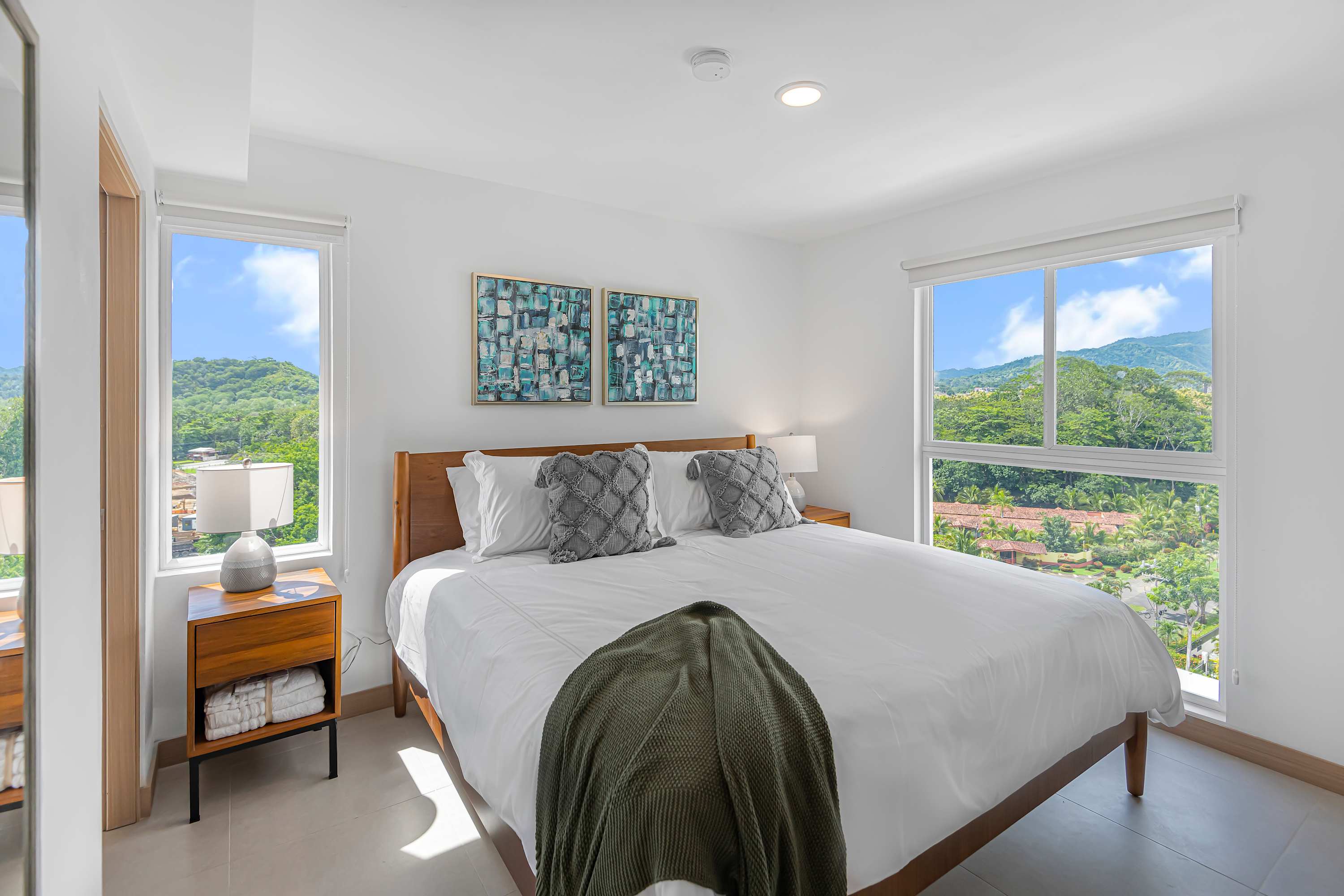 Serene bedroom featuring a wooden bed frame with white bedding and grey decorative pillows. Large windows on two sides provide abundant natural light and scenic views of green hills and distant buildings. Two abstract artworks adorn the wall above the bed.