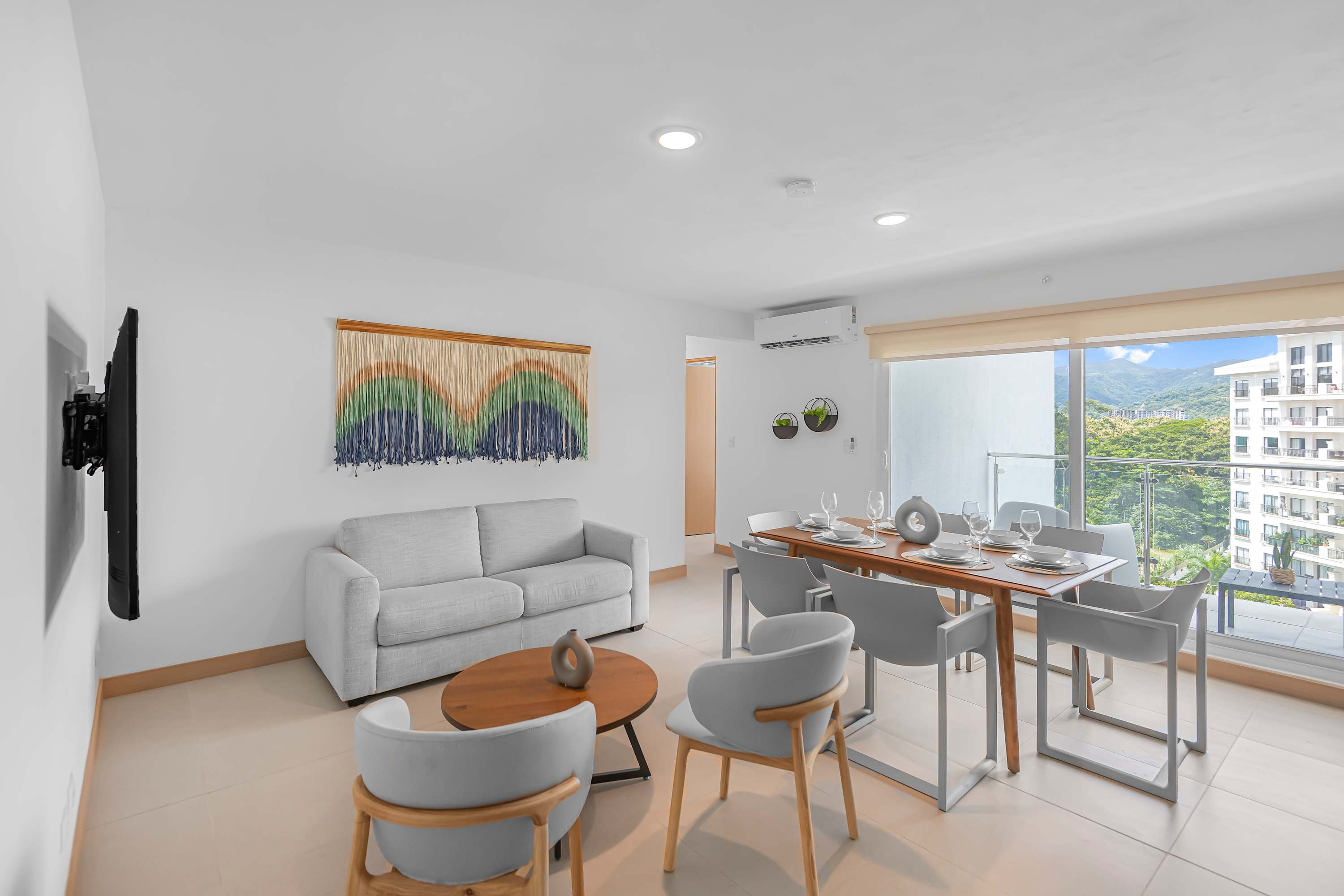 Spacious open-concept living and dining area with a light grey sofa, two armchairs, and a wooden coffee table. A rectangular dining table with six chairs is set for meals. A large wall hanging adds a decorative touch, and a sliding glass door leads to a balcony.