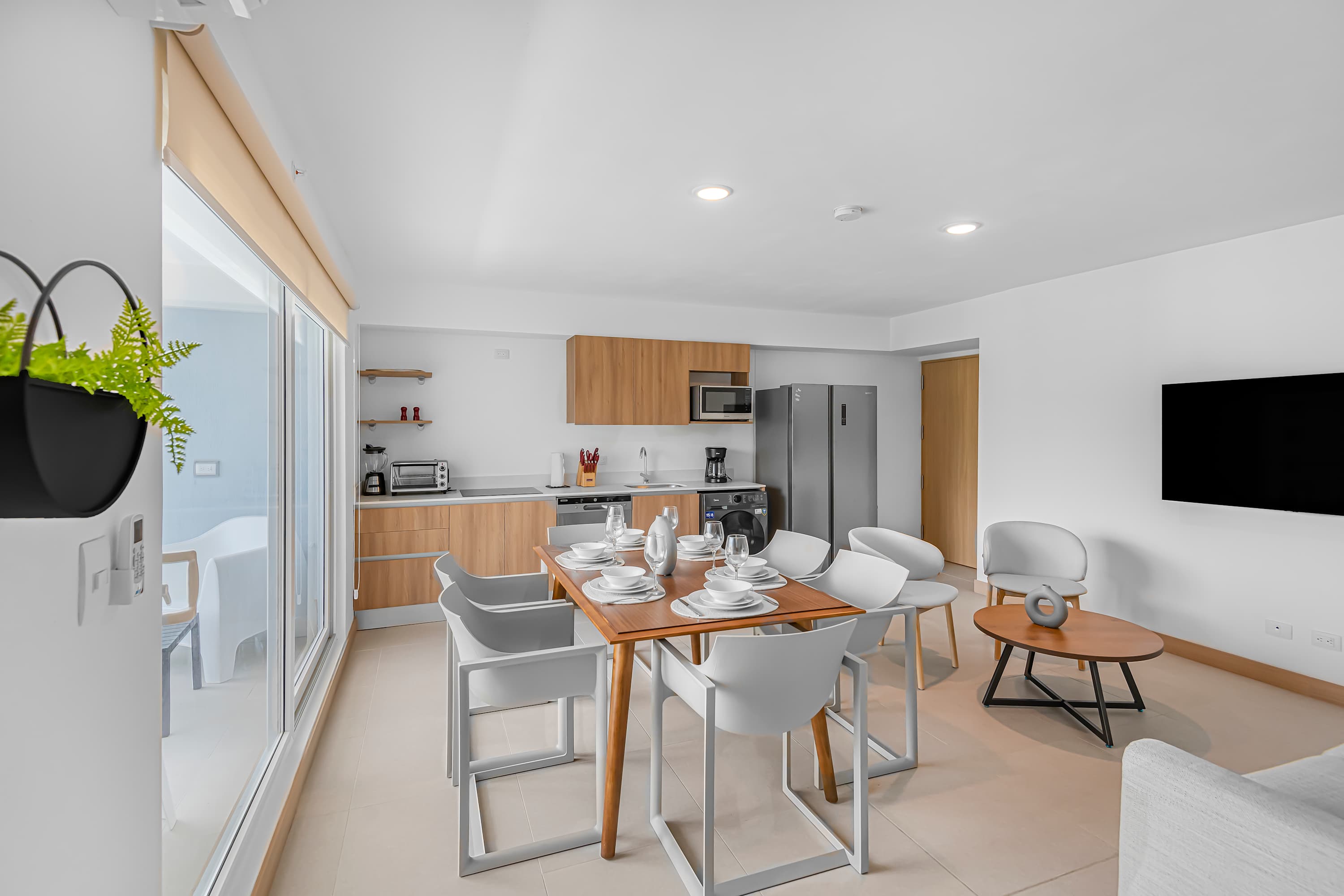 Bright open-plan living area with a dining table for six, a compact kitchen with light wood cabinets, and a living room with a light grey sofa. Large sliding glass doors open to a balcony, providing natural light and views of the exterior.
