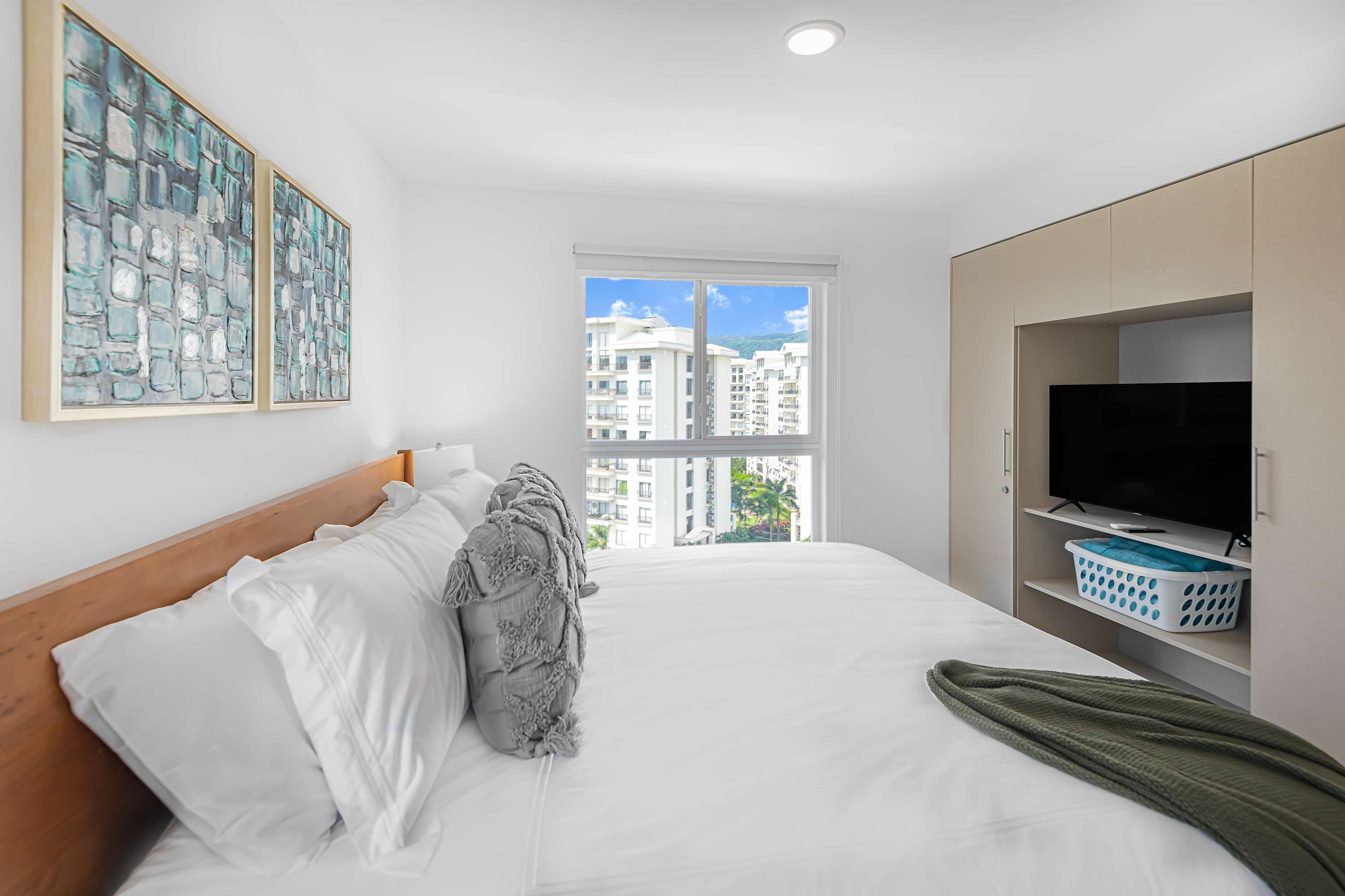 Cozy bedroom with a wooden bed frame, white bedding, and grey pillows. A large window offers views of nearby buildings. Built-in light wood wardrobes flank a recessed area with a television and shelving.