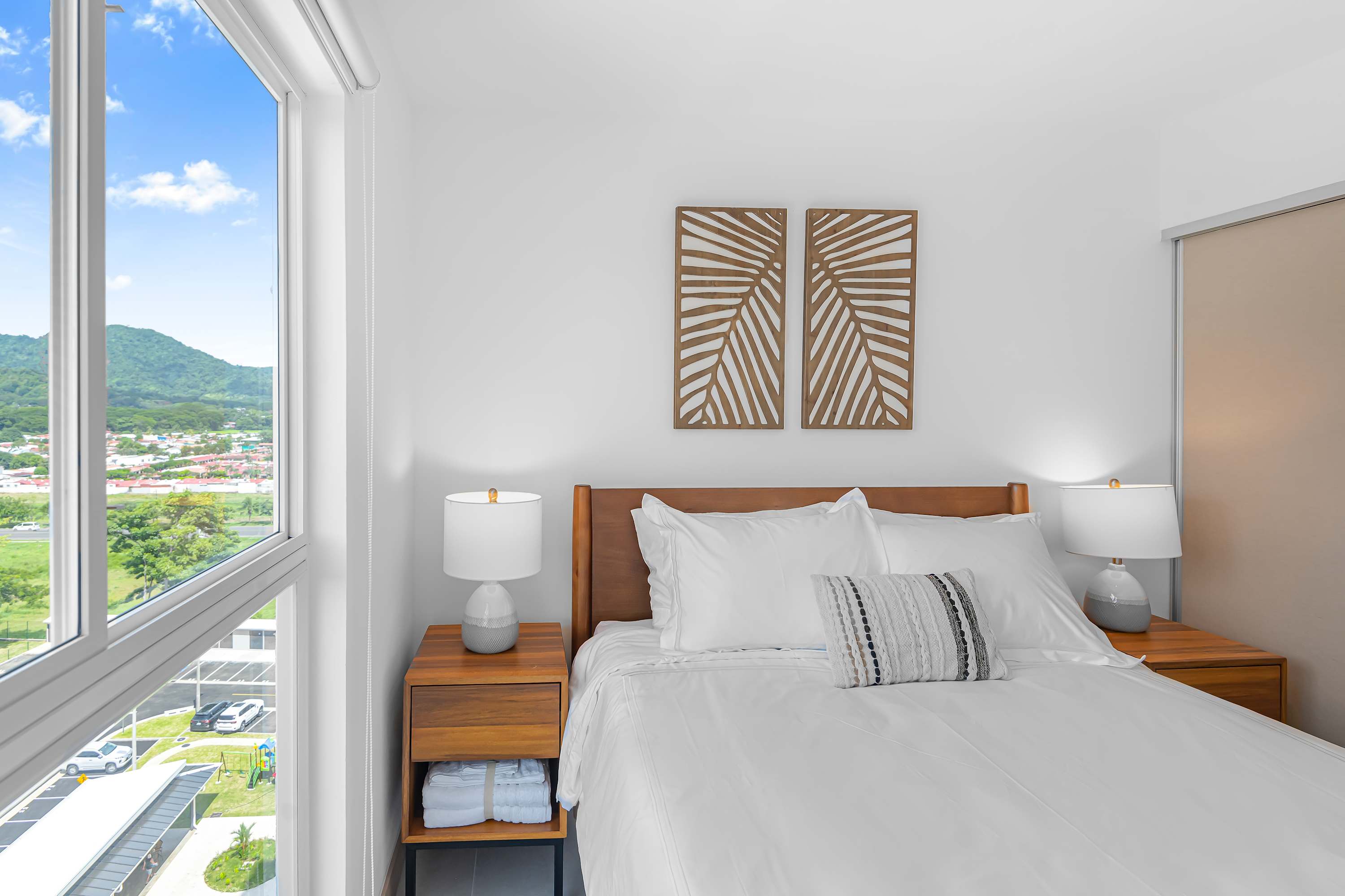Bright bedroom featuring a queen bed with white linens, a wooden headboard, and matching nightstands with lamps. Two decorative wooden panels adorn the wall above the bed, and a large window provides a scenic view of mountains and a town.