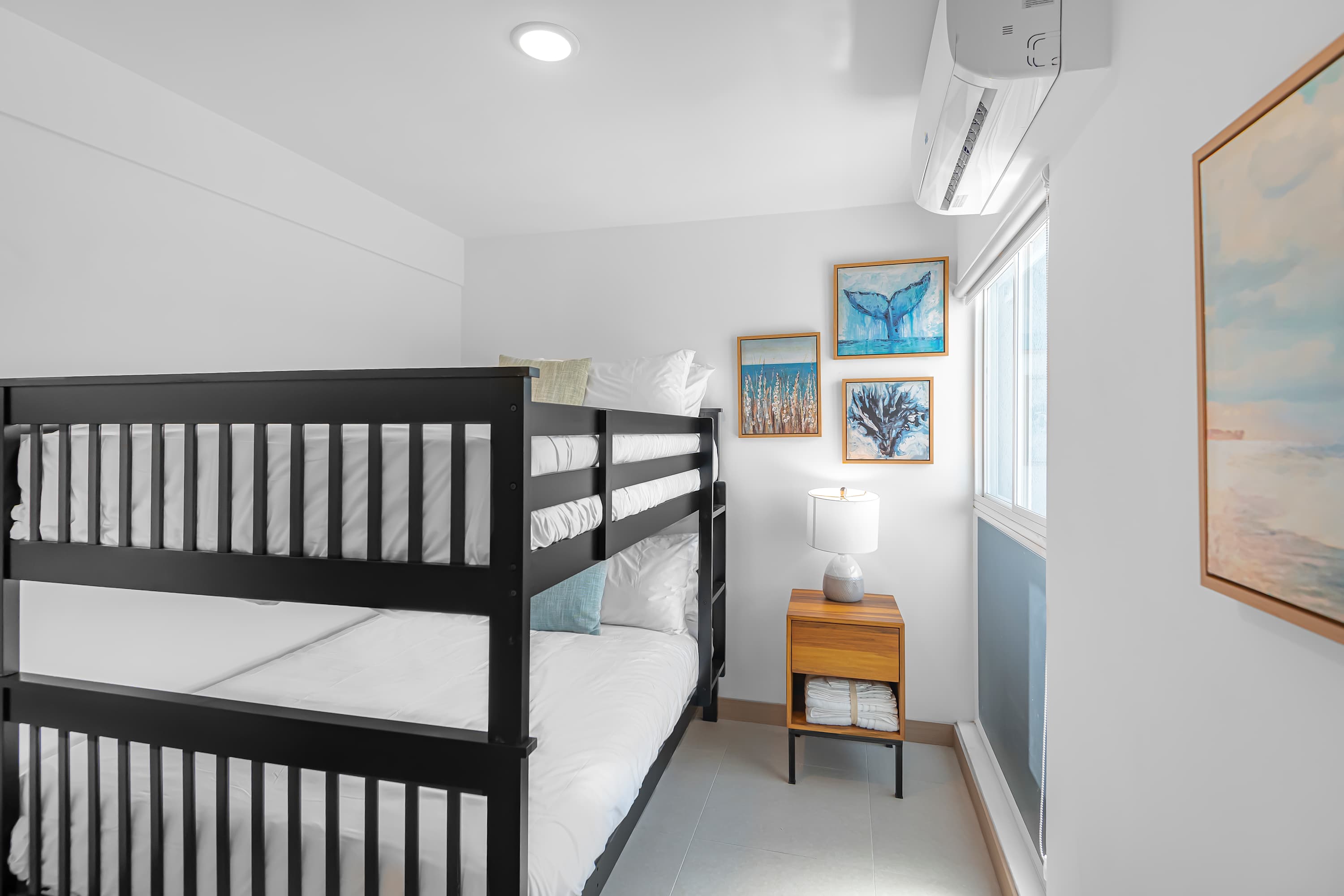 Modern bedroom featuring black bunk beds with white linens, a wooden nightstand with a lamp, and three framed artworks with marine themes on the wall. An air conditioning unit is mounted above the window.