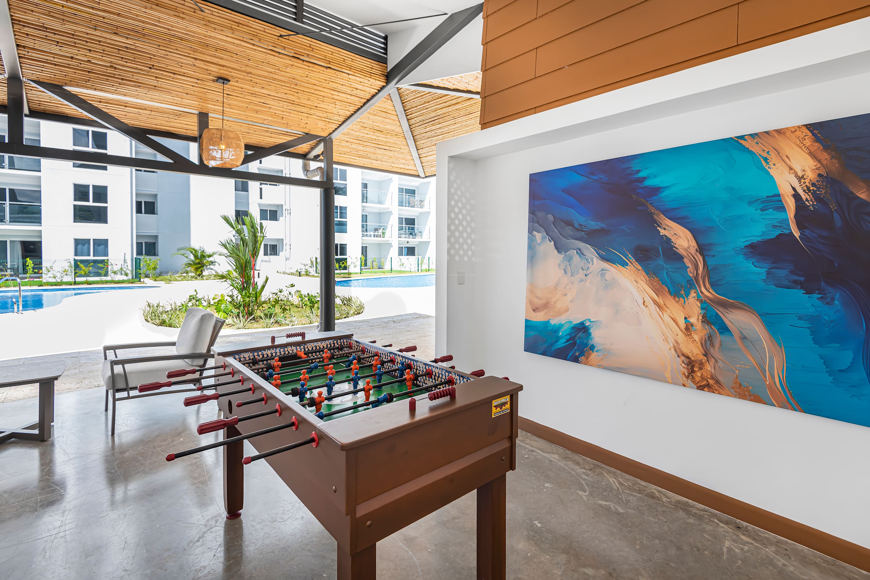 Covered outdoor recreation area featuring a foosball table, a large abstract painting on the wall, and views of the communal pool area. The ceiling has a natural wood slat design.