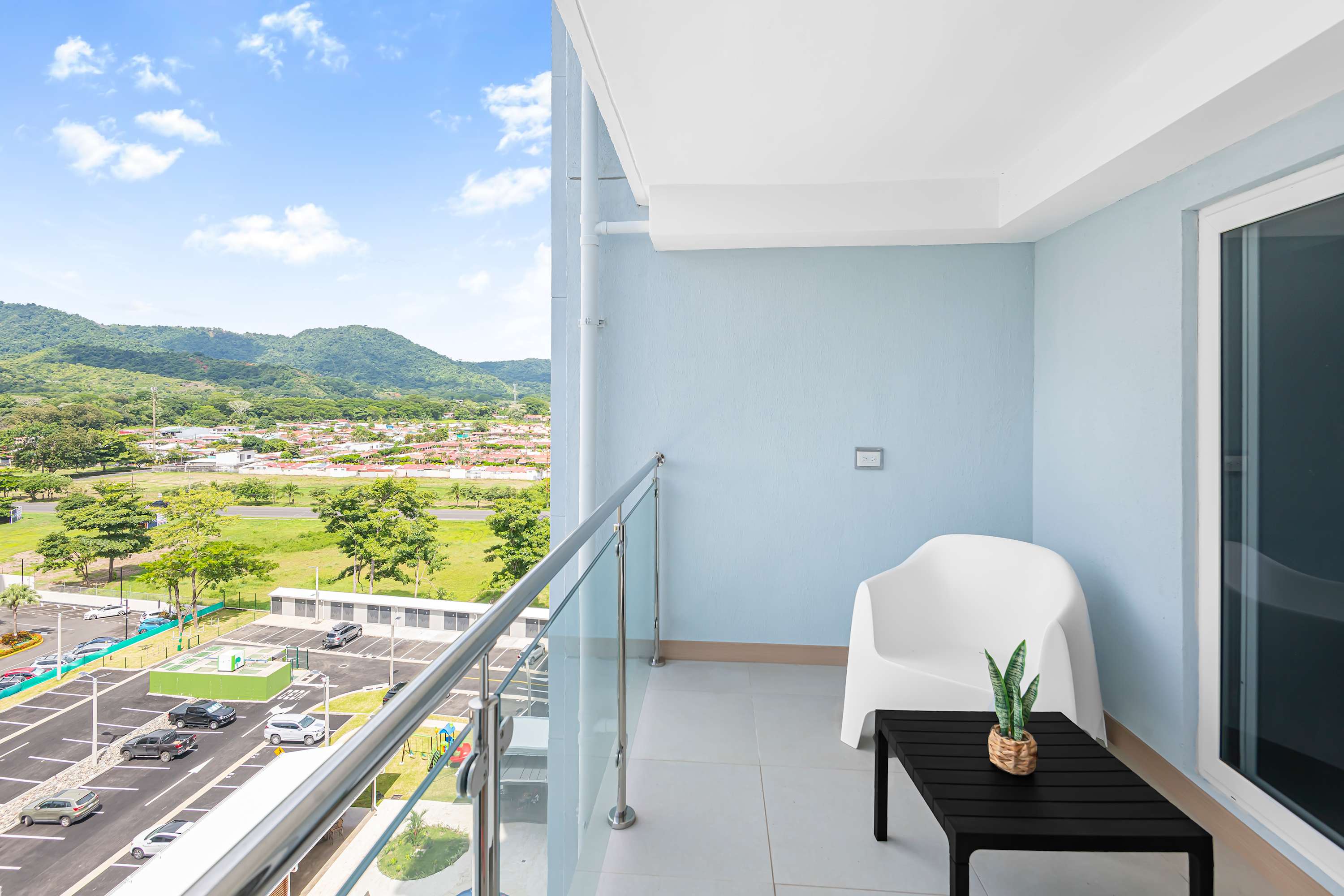Private balcony with light blue walls and a glass railing, offering expansive views of a valley with distant mountains and a parking lot below. A white modern chair and a small black table with a potted plant furnish the space.
