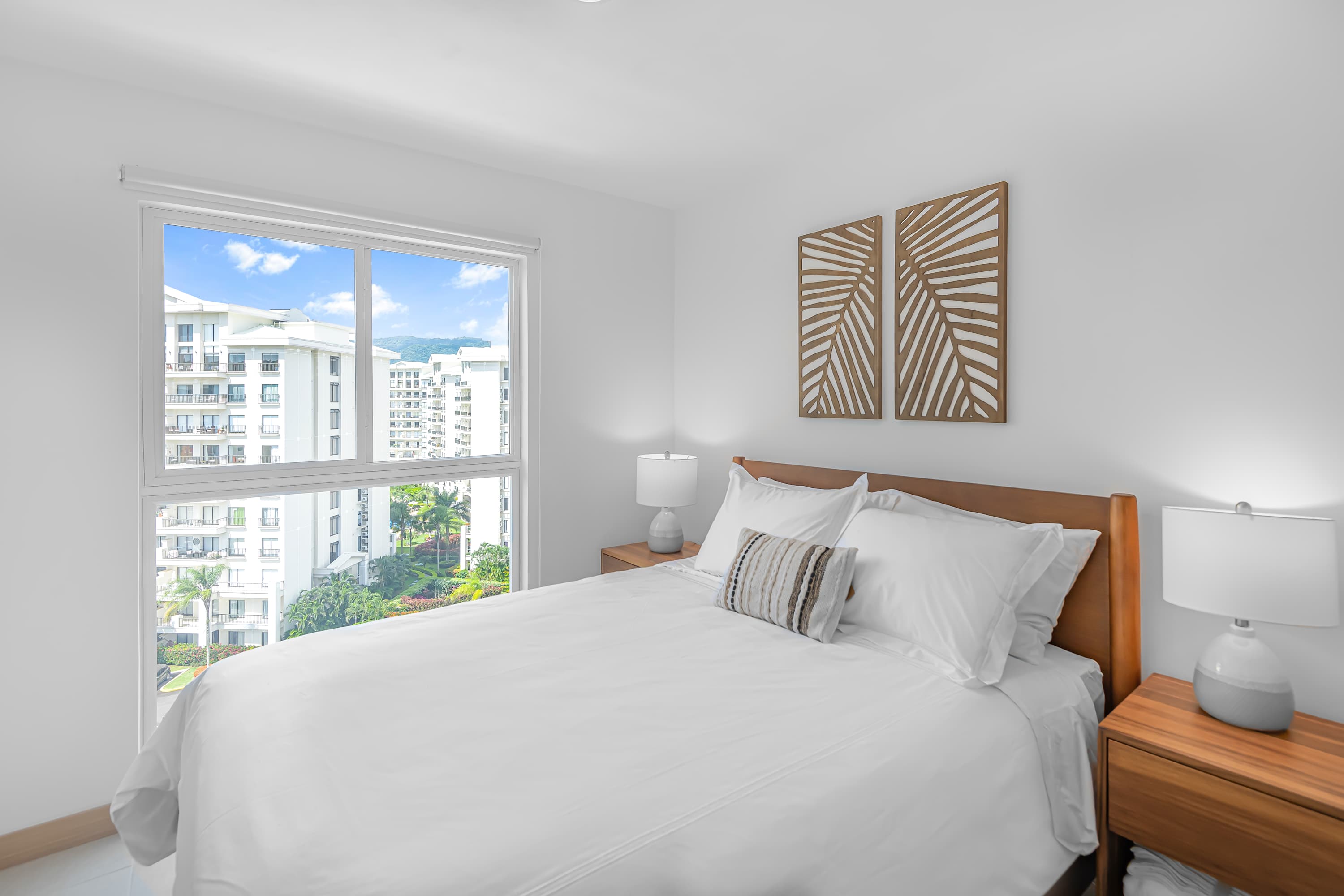 Modern bedroom with a queen-sized bed, crisp white bedding, a wooden headboard, two wooden nightstands with lamps, and two decorative wooden wall art pieces above the bed. A large window offers a view of other buildings and greenery.
