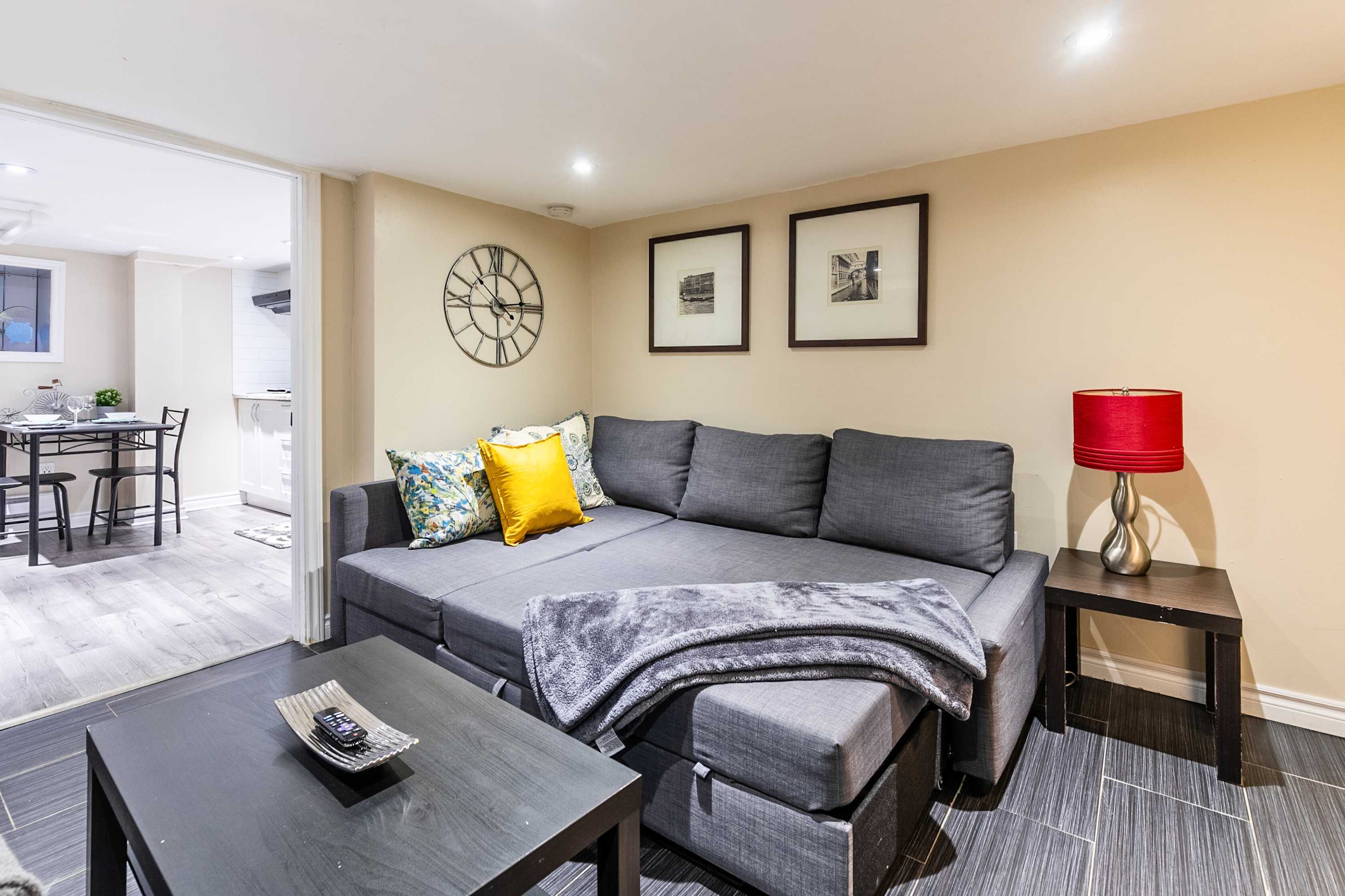 Cozy living room featuring a large grey sectional sofa with colorful throw pillows and a grey blanket, a dark wood coffee table, and a side table with a red lamp, all set on dark grey tiled flooring. A large wall clock and two framed pictures adorn the light beige wall.