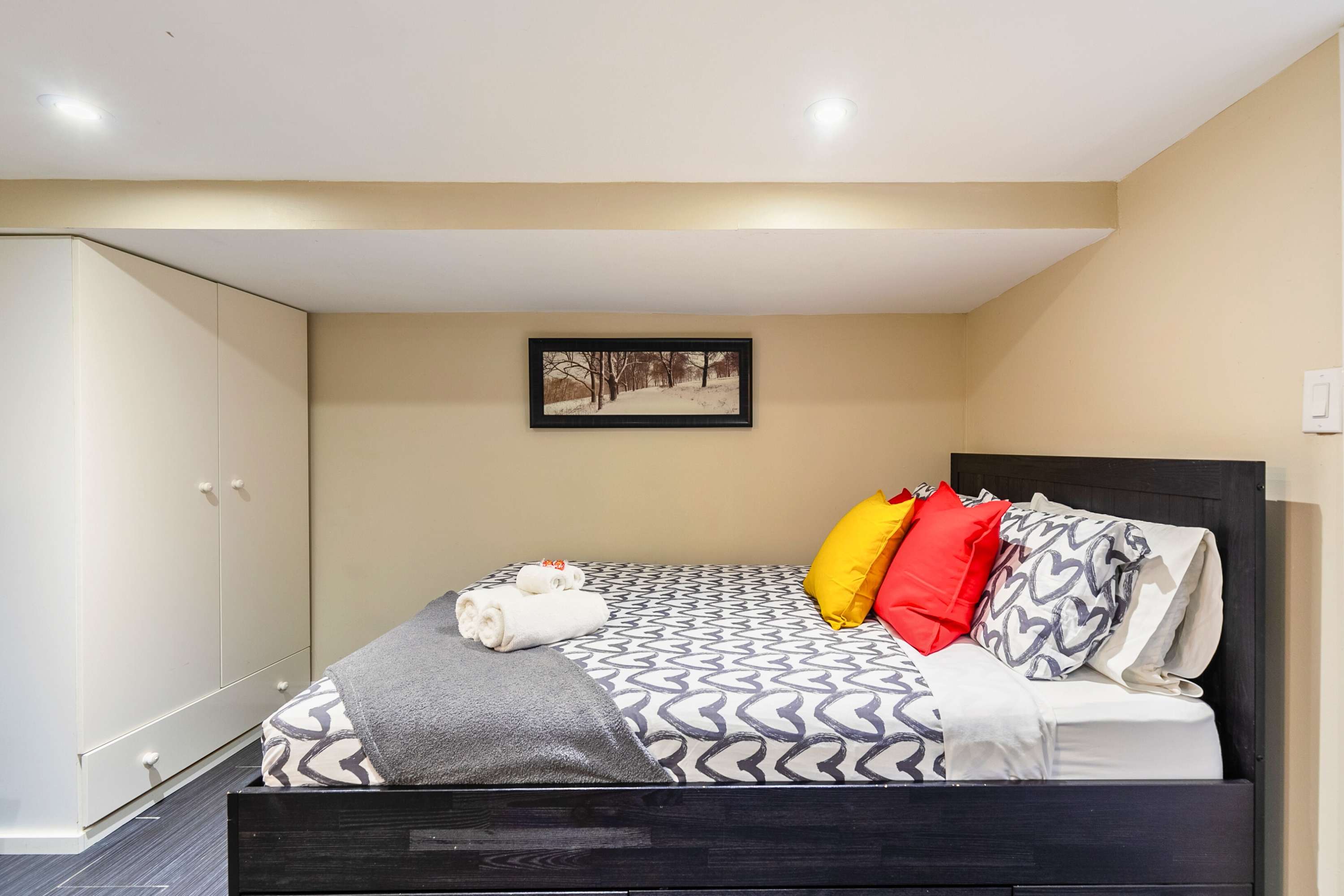 View of the bedroom showcasing a black bed with patterned bedding and colorful pillows, positioned against a light beige wall with a framed winter scene. Built-in white cabinets with round knobs line the left wall, complementing the dark grey tiled floor.