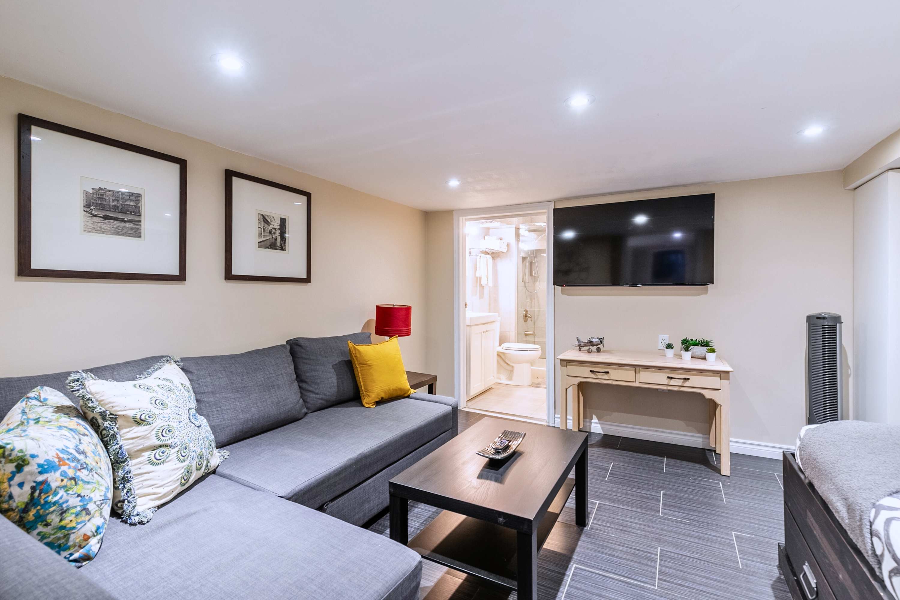 A comfortable living room with a gray L-shaped sectional sofa, a dark coffee table, and a wall-mounted flat-screen TV. Dark tiled flooring and light beige walls create a warm ambiance, with a glimpse of the bathroom through an open doorway.
